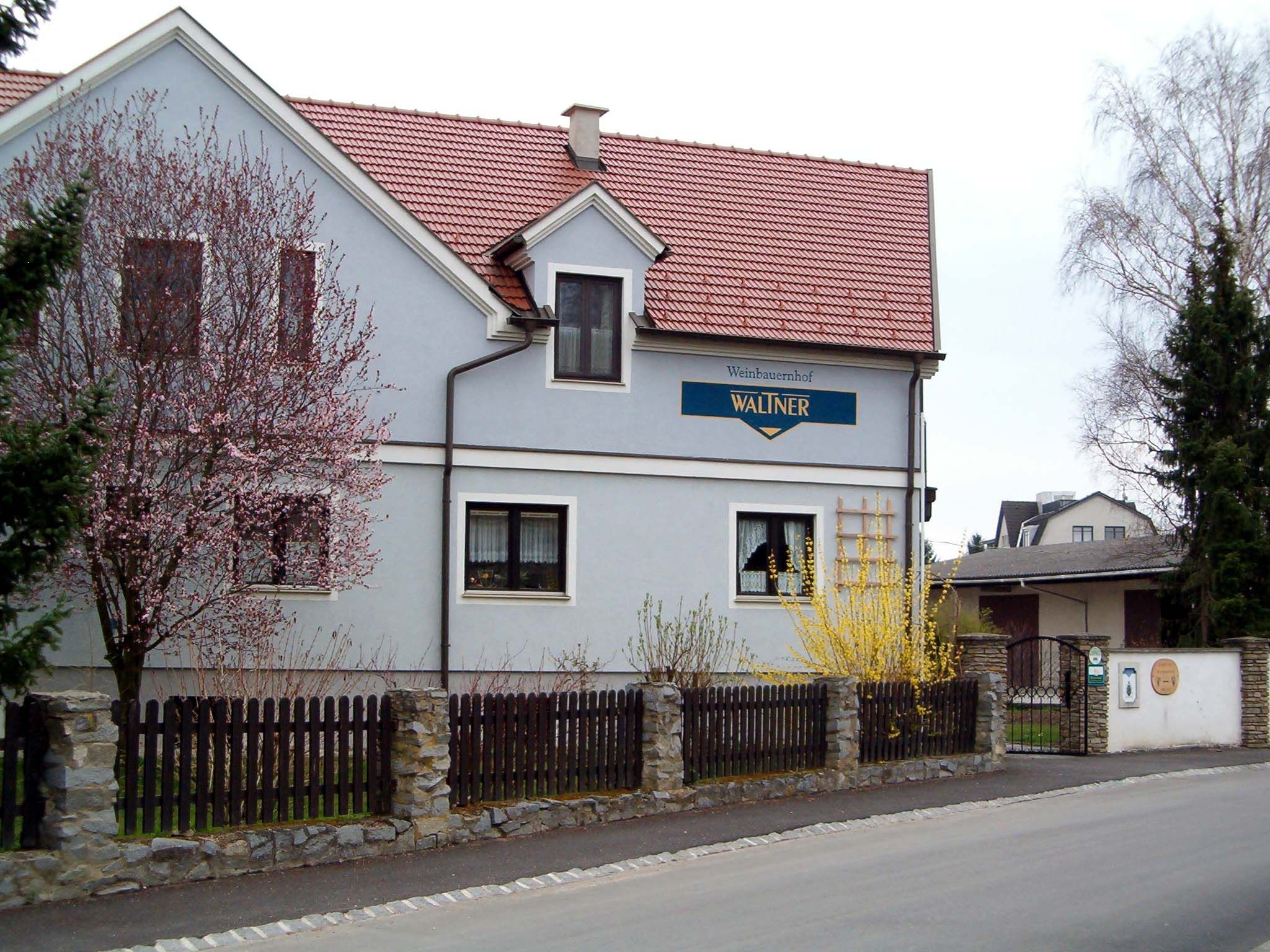 Ein zweistöckiges Haus mit rotem Dach und blauer Fassade, umgeben von blühenden Bäumen und einem Holzzaun.