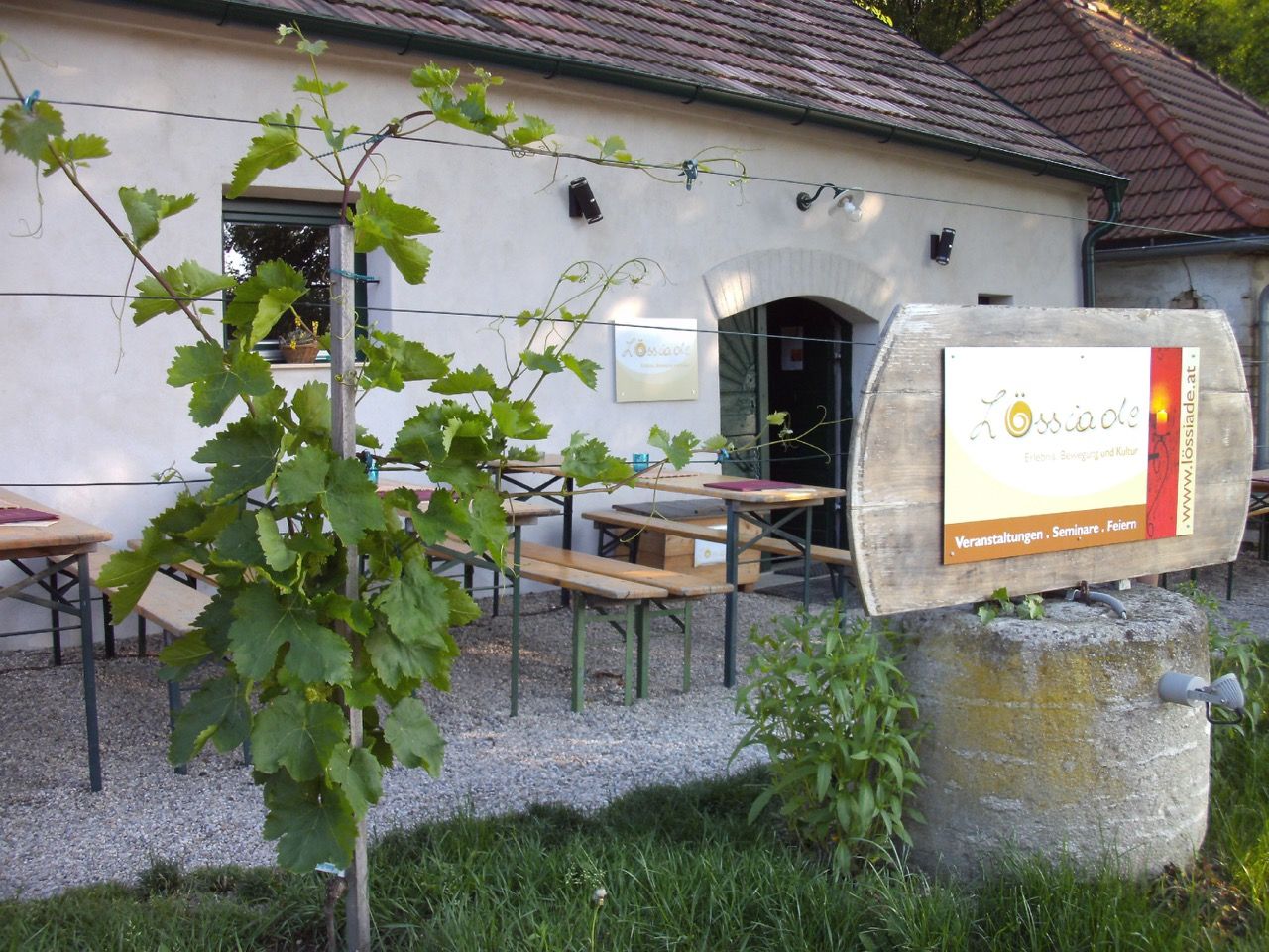 Gastgarten der Lössiade mit Holzbänken, Weinreben im Vordergrund und Fass-Schild vor einem traditionellen Gebäude bei Tageslicht.