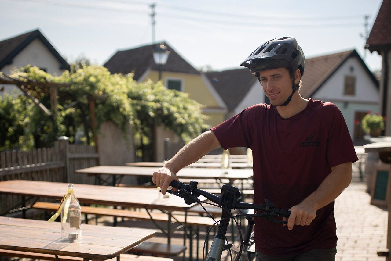 Ein junger Radfahrer genießt die warmen Sonnenstrahlen, während er durch die charmante Kellergasse Oagossn radelt. Umgeben von malerischen Weinbergen und rustikalen Holzhütten, vermittelt die Szenerie ein Gefühl von Freiheit und Abenteuer. Hier in der idyllischen Landschaft des Weinviertels wird jeder Moment zum unvergesslichen Erlebnis.