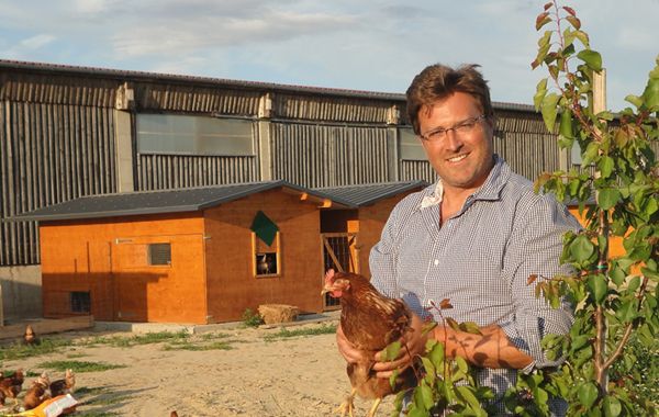 Ein Mann steht mit einem Huhn in der Hand auf einem Bauernhof.