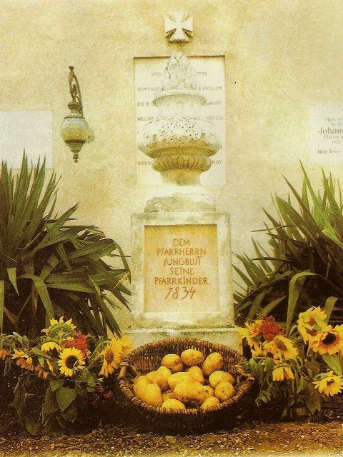 Denkmal mit Inschrift, umgeben von Pflanzen und einem Korb mit Kartoffeln und Sonnenblumen.