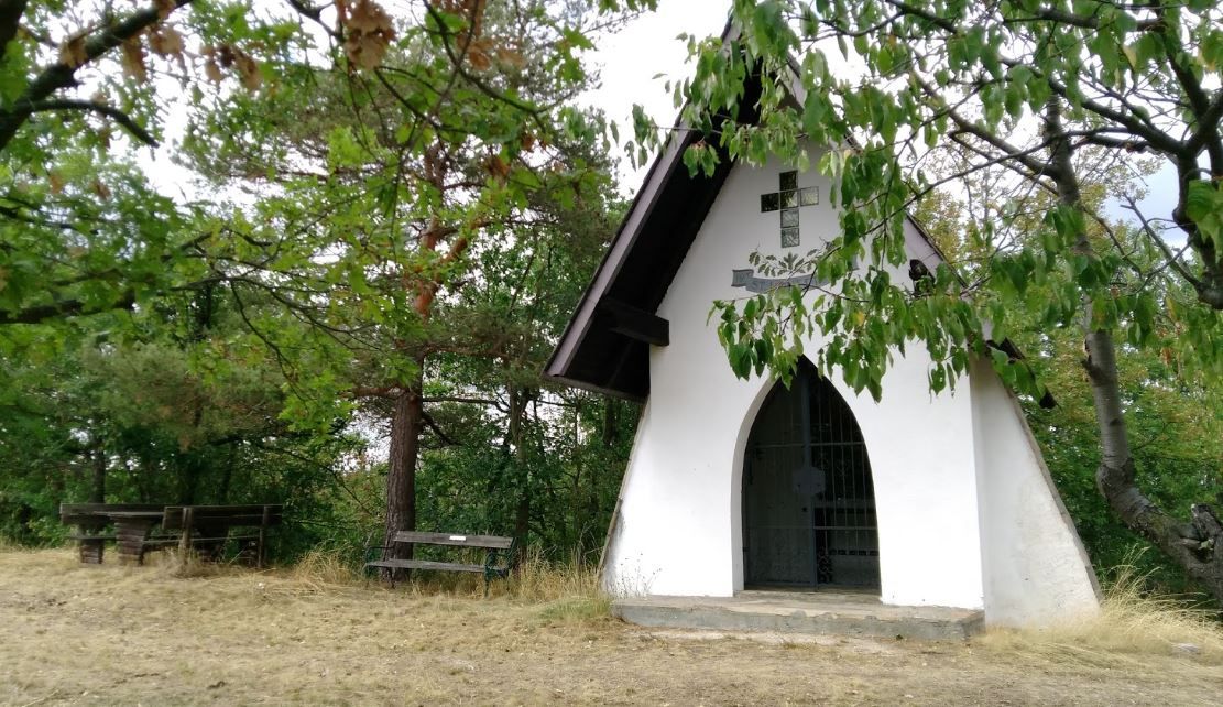 Kleine weiße Kapelle im Wald mit Holzbänken davor.