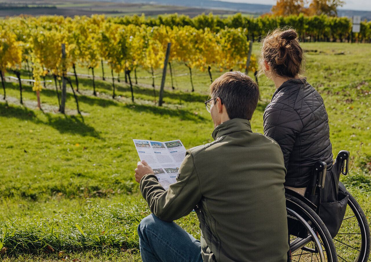 Eine junge Frau im Rollstuhl und ein Mann genießen den Ausblick auf eine mit herbstlich gefärbten Weingärten durchzogene Landschaft. Der Mann hält einen Folder mit Tipps in der Hand.