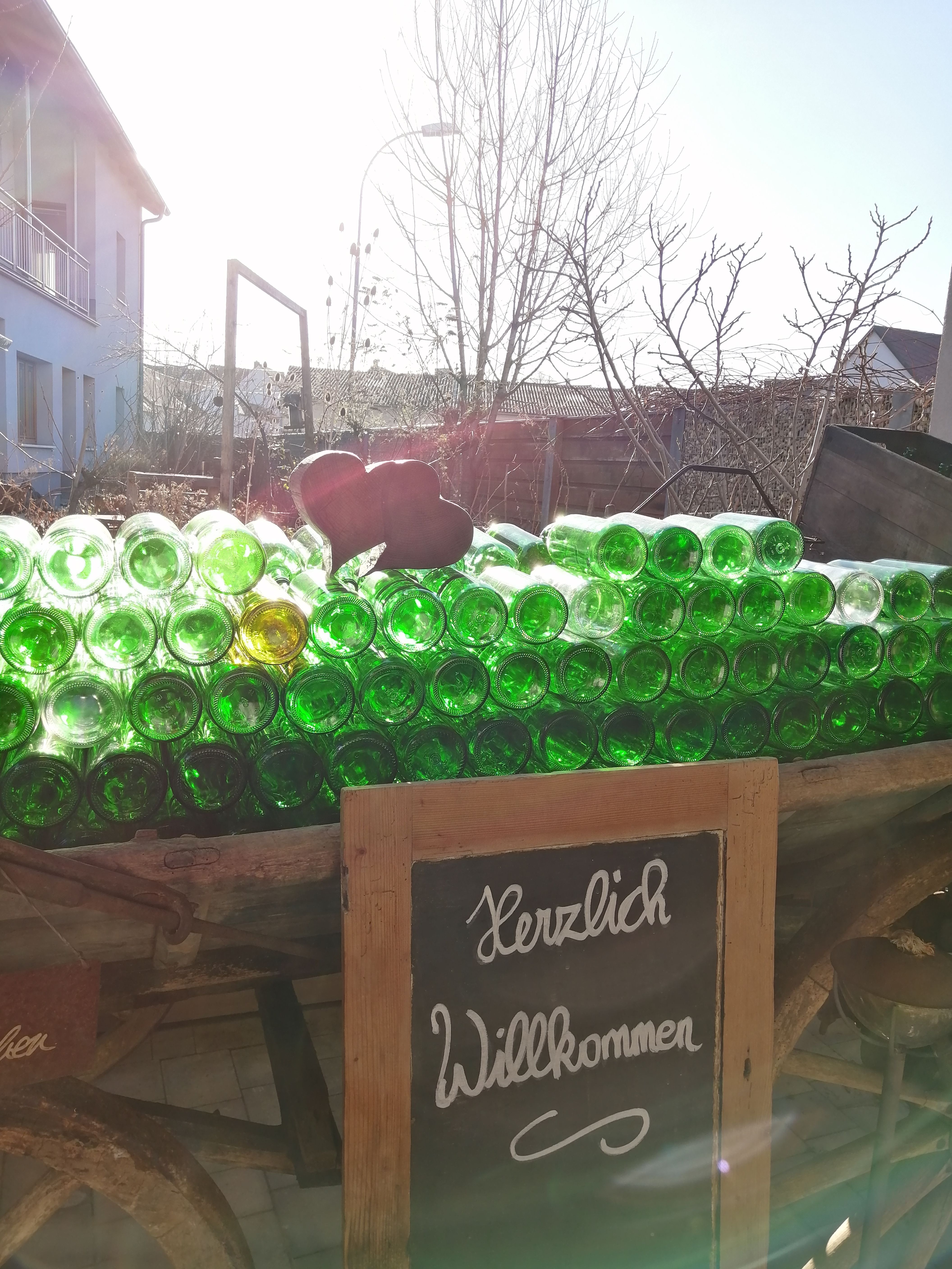 Ein Stapel grüner Glasflaschen auf einem Wagen mit einem Schild 'Herzlich Willkommen' davor, im Hintergrund ein Haus und Bäume.