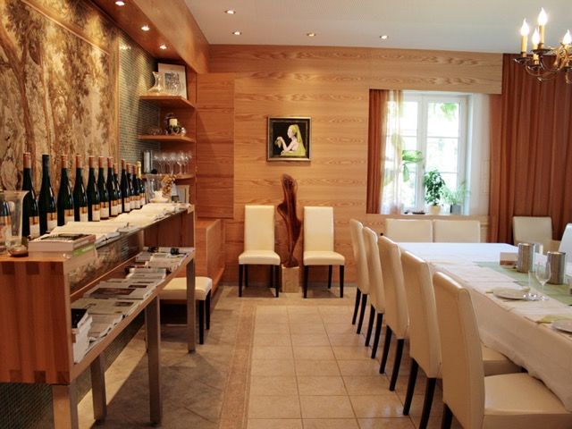 Ein Verkostungsraum mit Holzwänden, einem langen Tisch mit weißen Stühlen und Weinflaschen auf einem Regal.