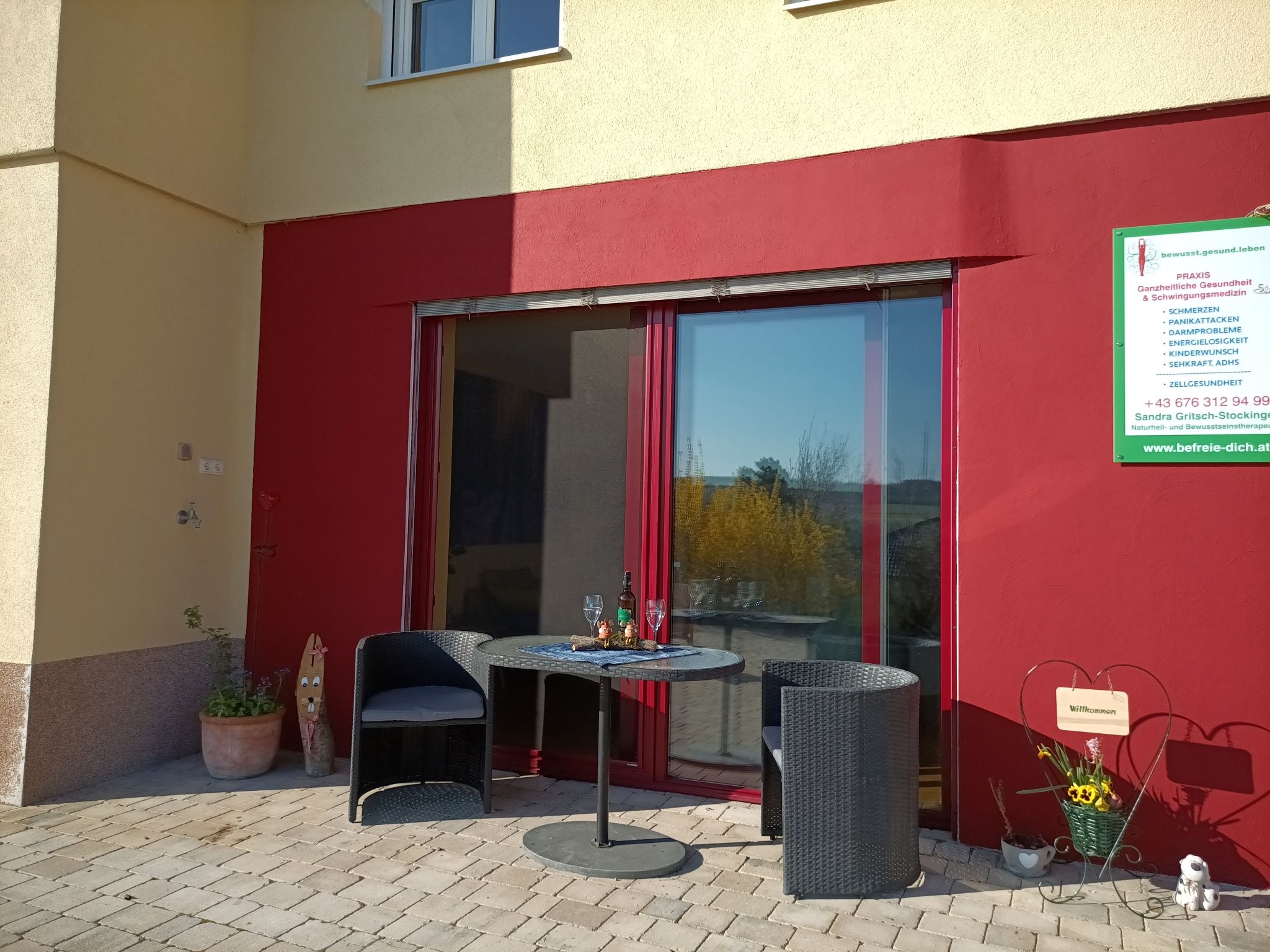 Terrasse mit zwei Stühlen und einem Tisch vor einer roten Wand, daneben ein Schild mit Informationen.