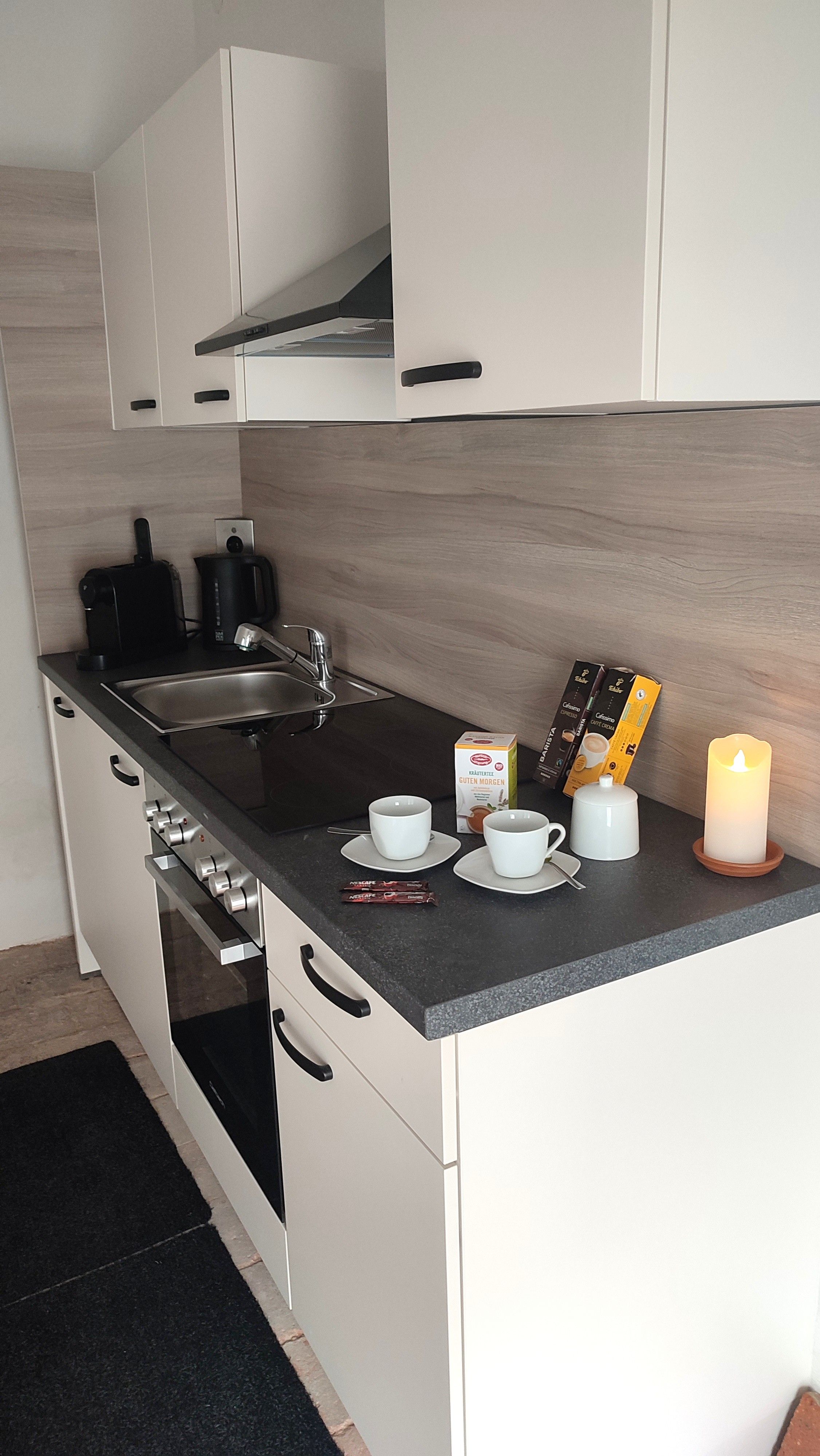 Kleine, moderne Küchenzeile mit weißen Schränken, Spüle, Herd und Kaffeemaschine. Auf der Arbeitsplatte stehen Tassen, eine Kerze und Kaffeezubehör.