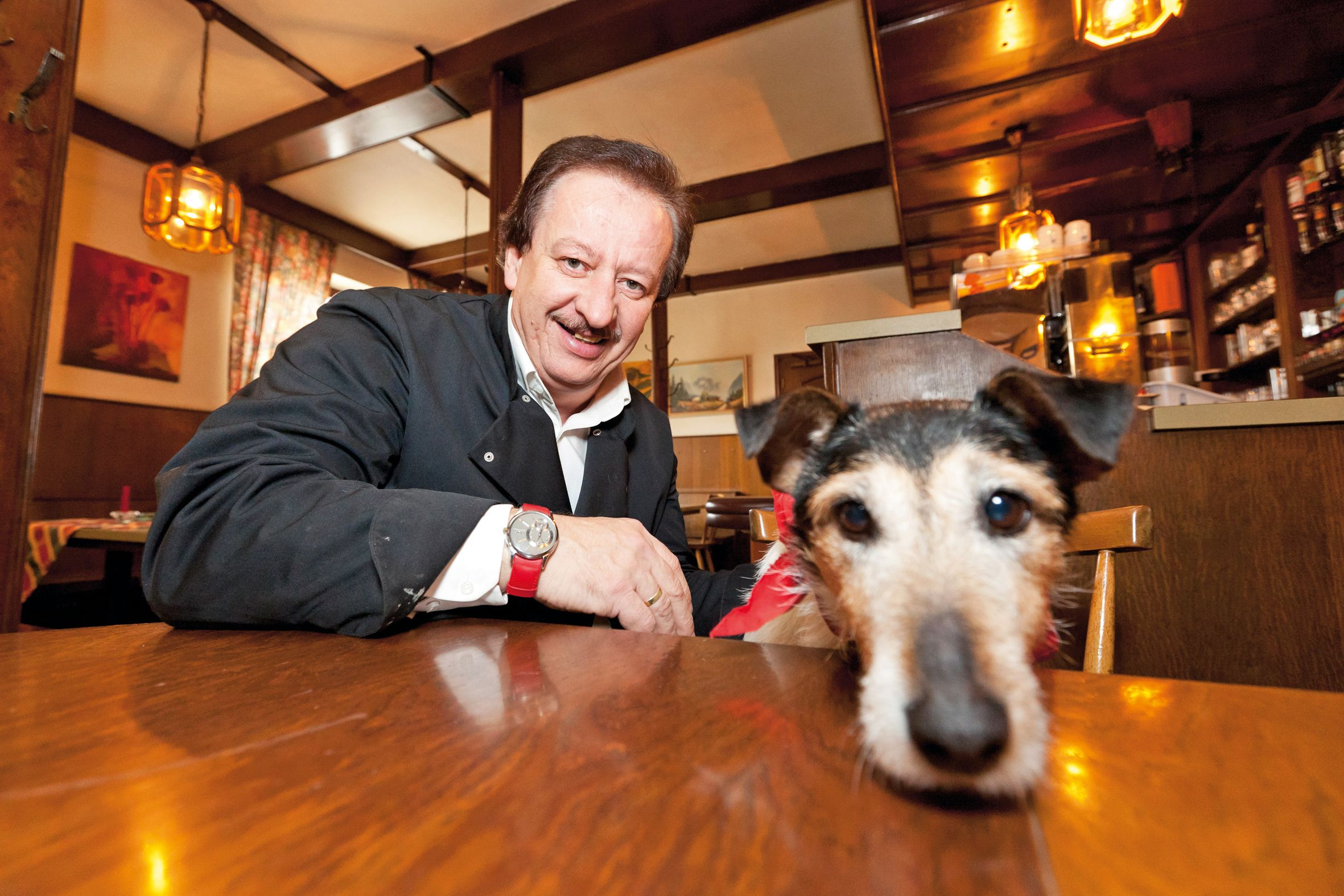 Ein Mann in einem Gasthaus lächelt, während ein Hund mit rotem Halstuch auf dem Tisch liegt.
