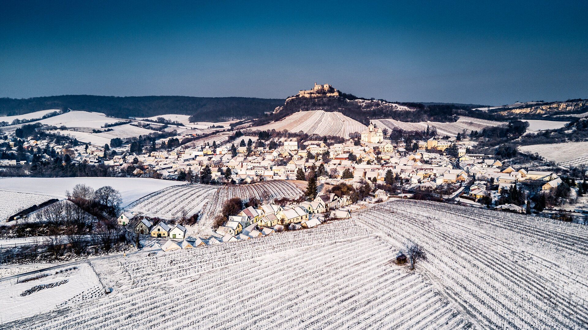In der winterlichen Kellergasse von Falkenstein erstreckt sich eine malerische Landschaft, die von einer sanften Schneedecke bedeckt ist. Die charmanten Häuser und die sanften Hügel schaffen eine friedliche Atmosphäre, die zum Verweilen einlädt. Hier, wo die Natur und die Tradition harmonisch verschmelzen, erleben Besucher die stille Schönheit des Winters.