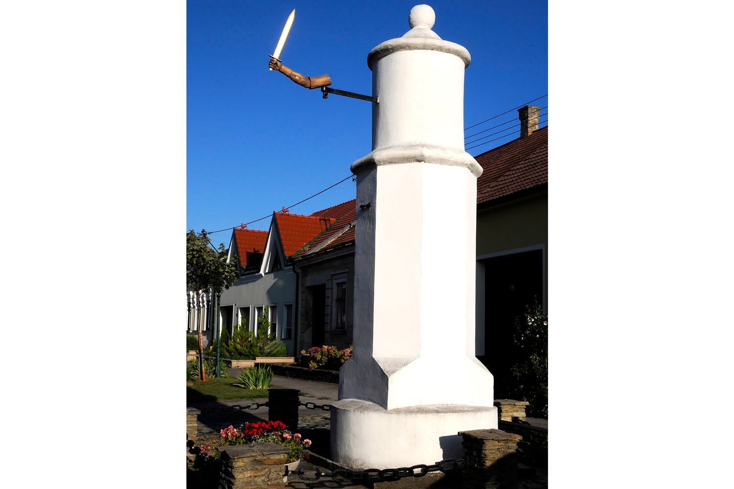 Ein weißer Pranger mit einer Holzhand, die ein Schwert hält, vor einem Haus mit roten Dächern.