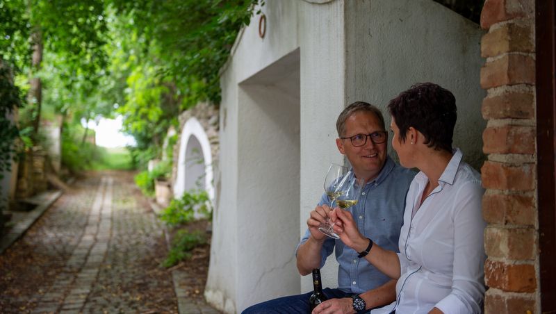 A couple sits in a wine cellar lane and clinks glasses of wine.