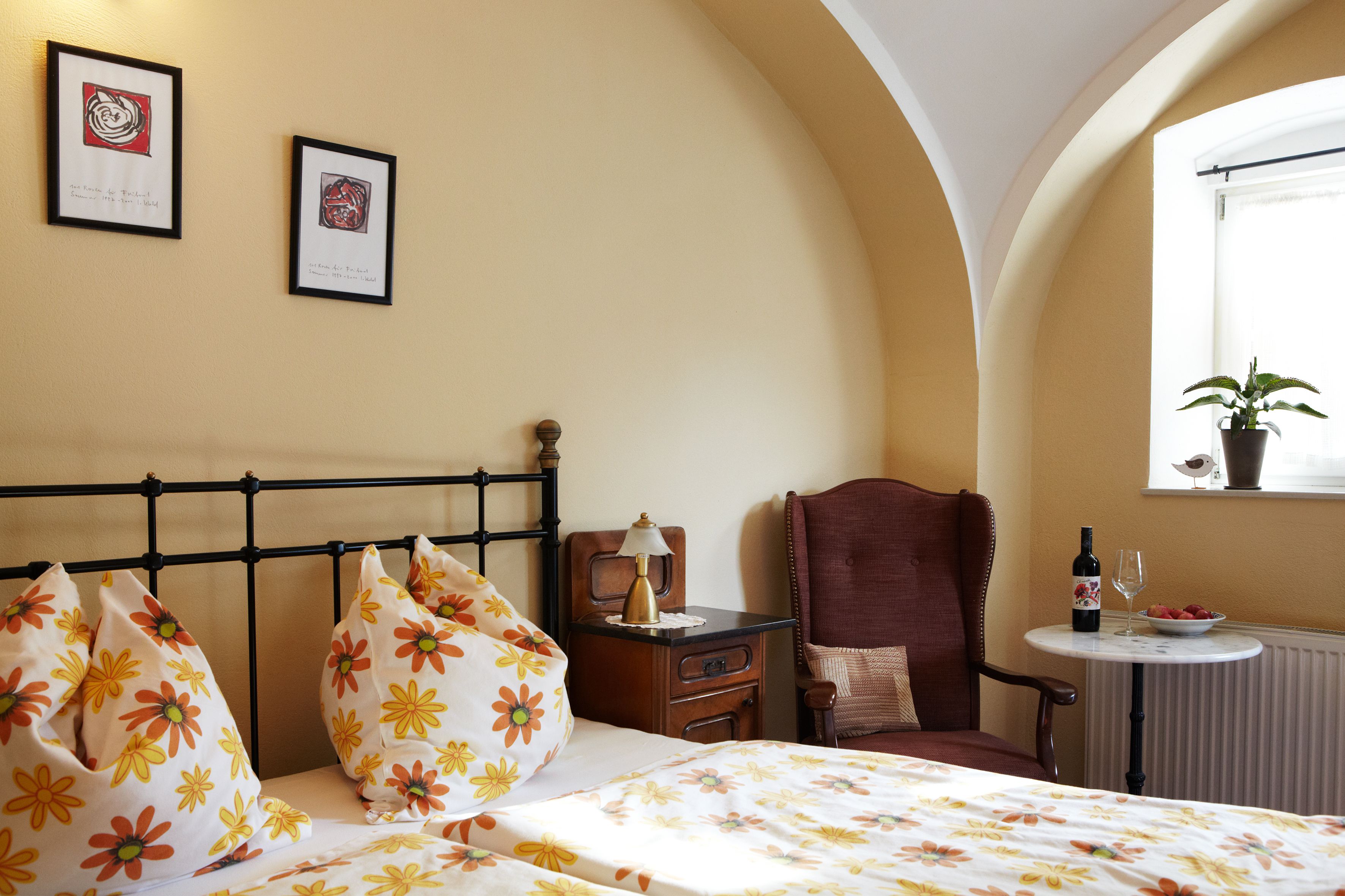 Gemütliches Gästezimmer mit Blumenbettwäsche, Sessel, Tisch mit Wein und Obst, und Pflanzen auf der Fensterbank.