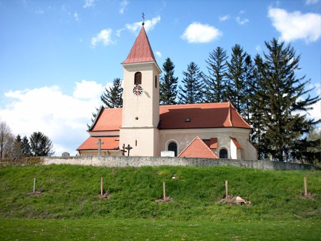 Eine Kirche mit rotem Dach und Turm vor einem Hintergrund aus Bäumen und blauem Himmel.
