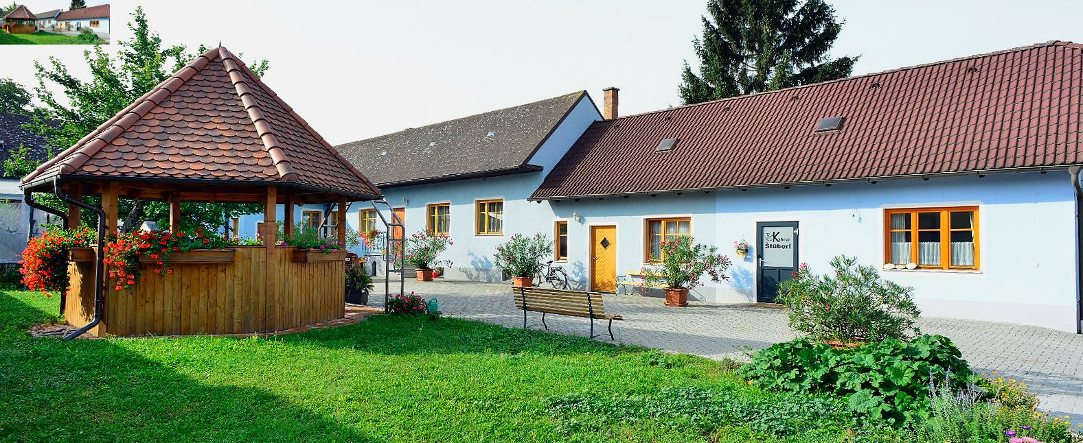 Ein idyllischer Hof mit einem Pavillon und einem blauen Gebäude mit roten Dächern, umgeben von Pflanzen.