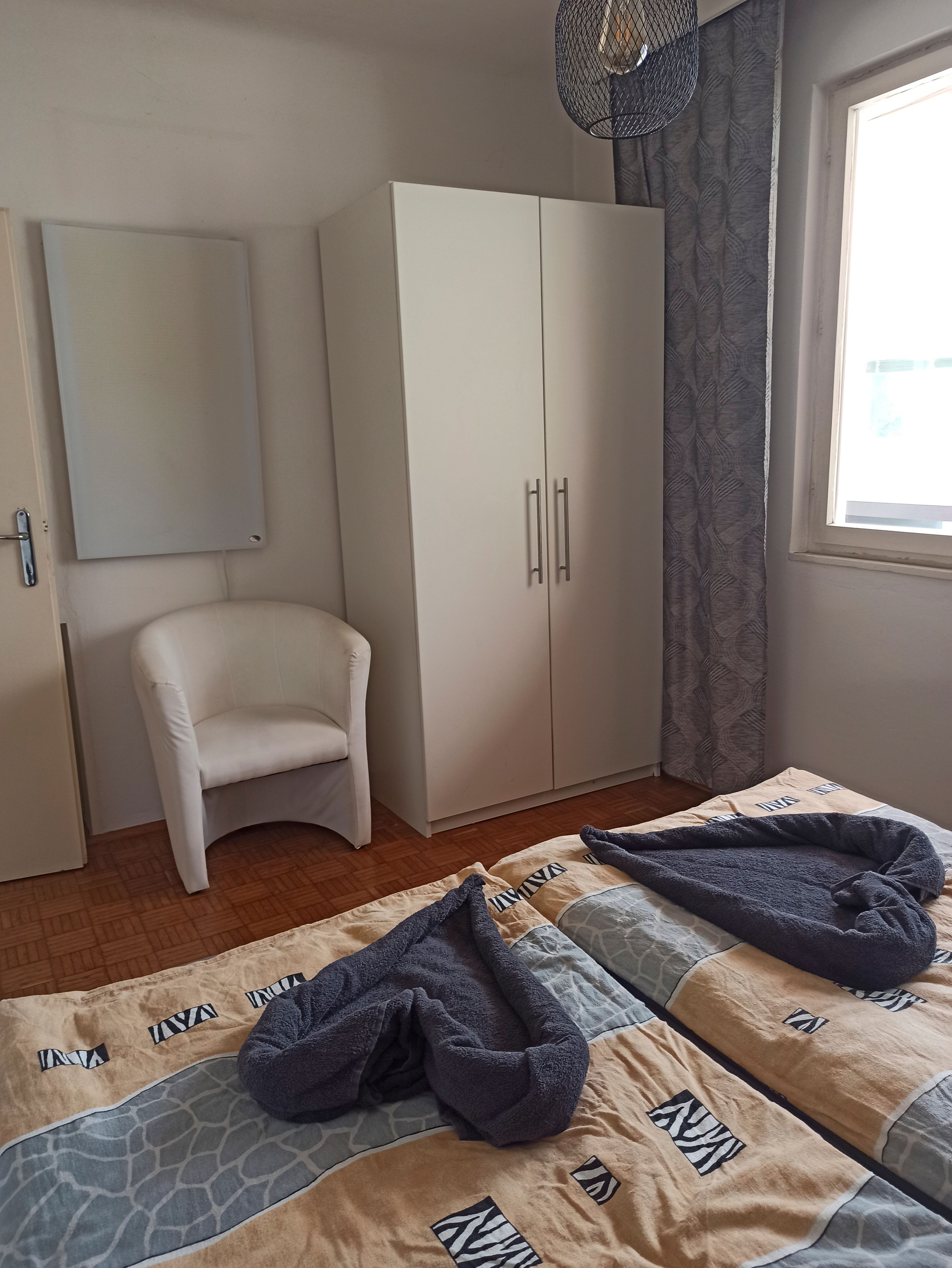 Ein Gästezimmer mit zwei Betten, einem weißen Schrank, einem Sessel und einem Fenster mit Vorhang.