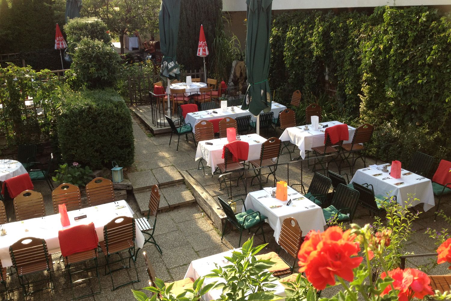 Ein gemütlicher Gastgarten mit gedeckten Tischen, umgeben von grünen Pflanzen und roten Blumen.