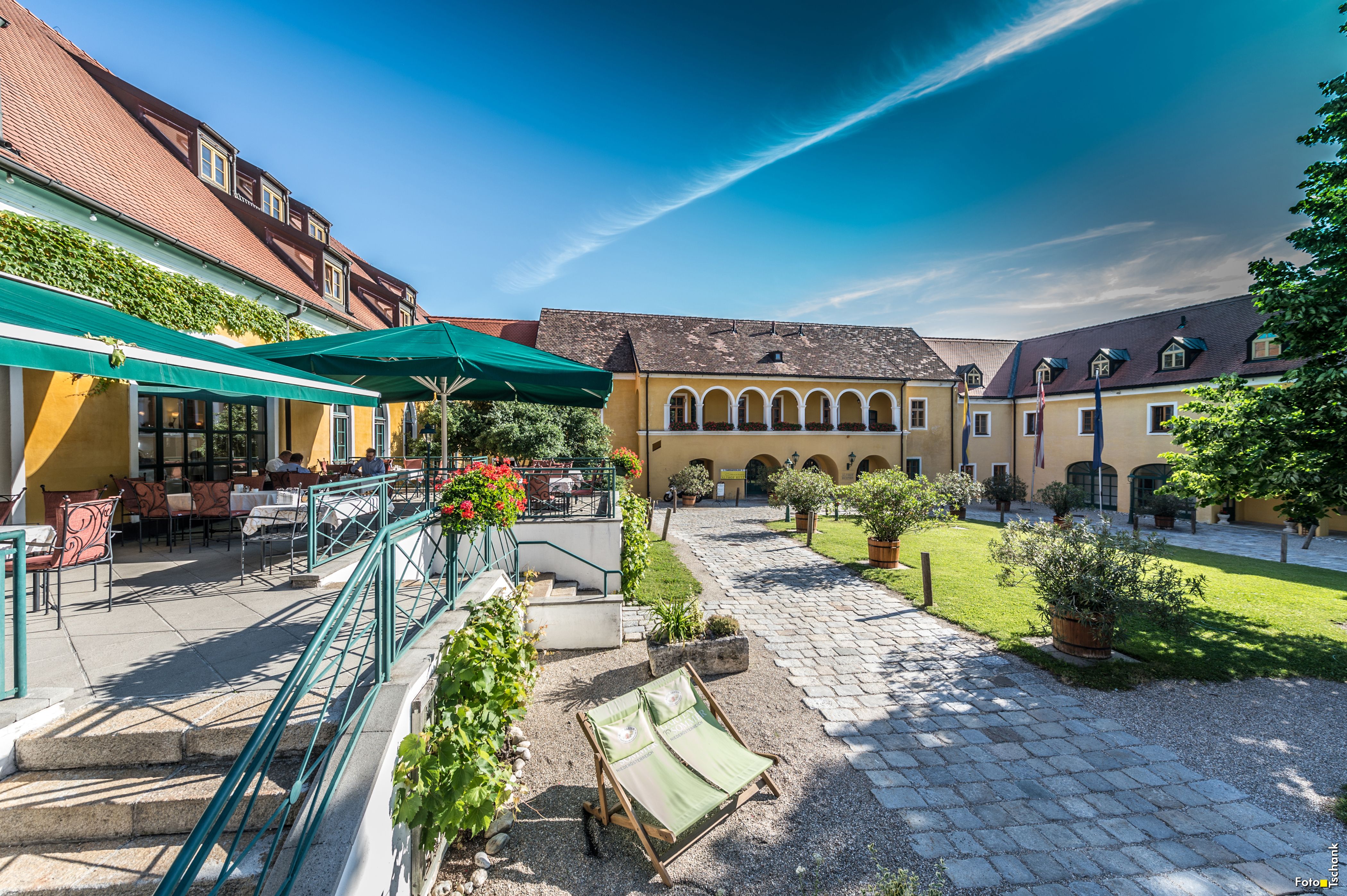 Ein sonniger Innenhof mit gelben Gebäuden, Terrassenbereich und Liegestühlen.