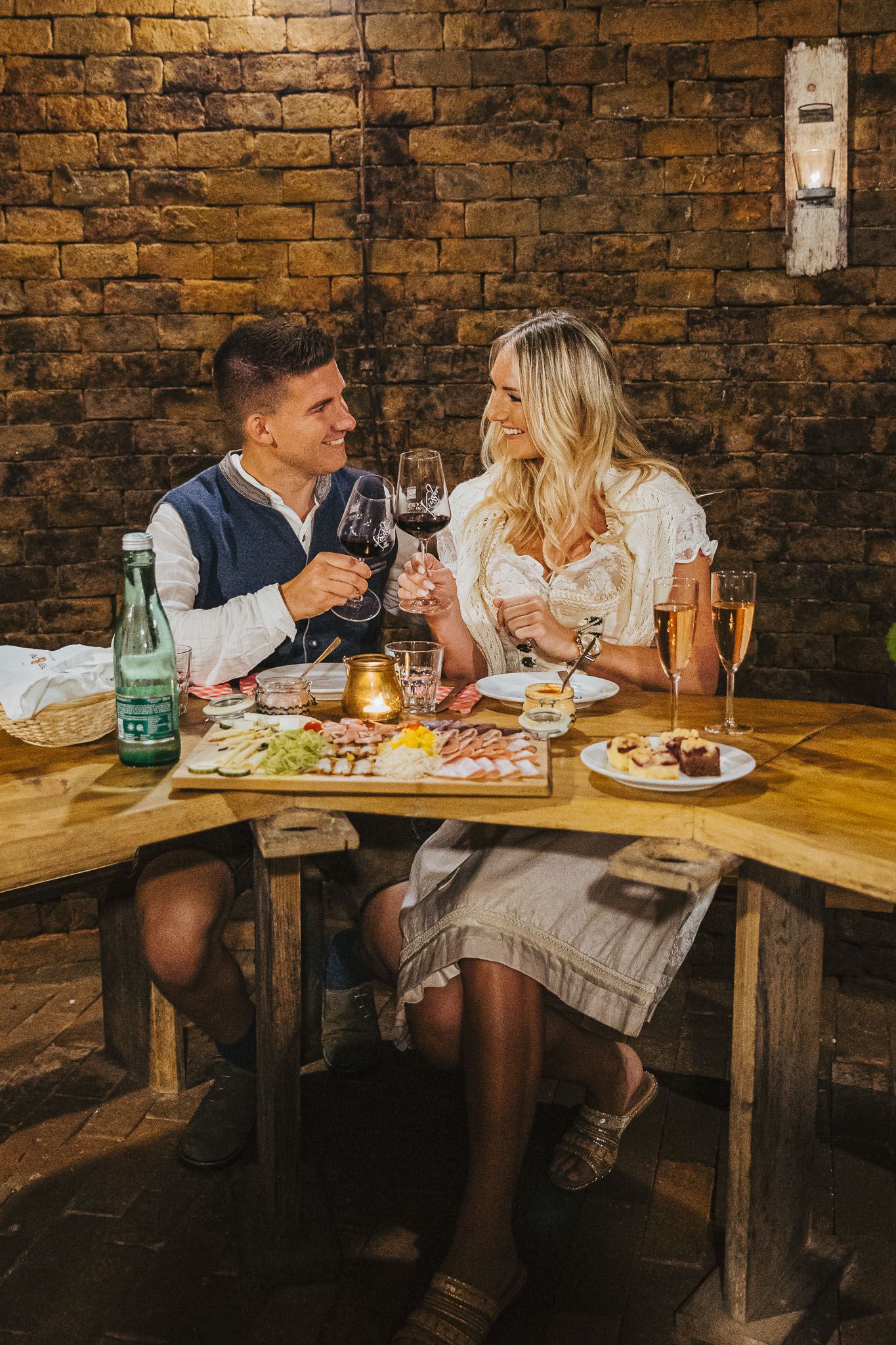 Paar beim Anstoßen mit Rotwein im Winzerhof Küssler an einem rustikalen Holztisch mit Jause und Desserts vor einer Ziegelwand.