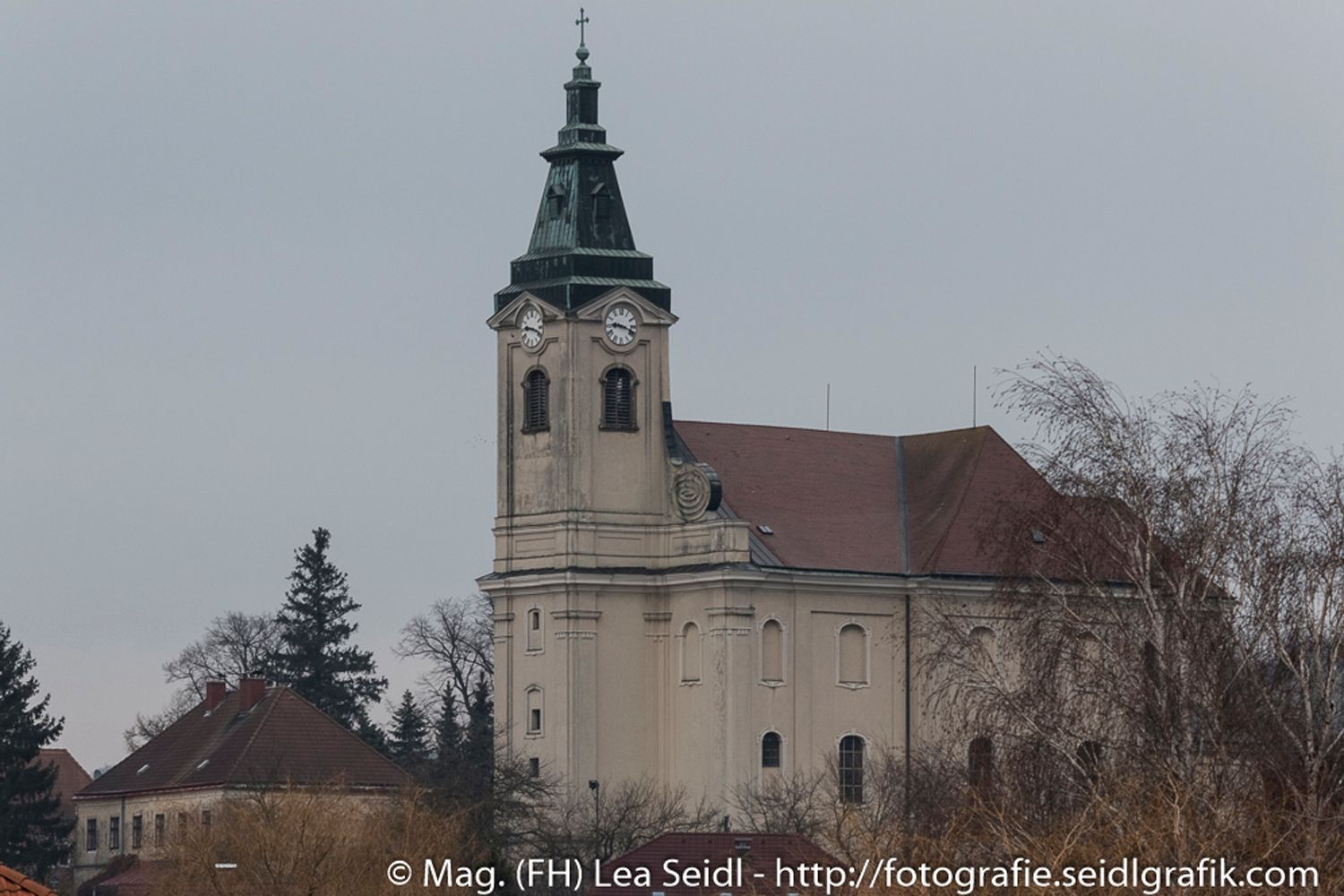Kirche in Niederhollabrunn mit Turm und Uhr, umgeben von Bäumen.