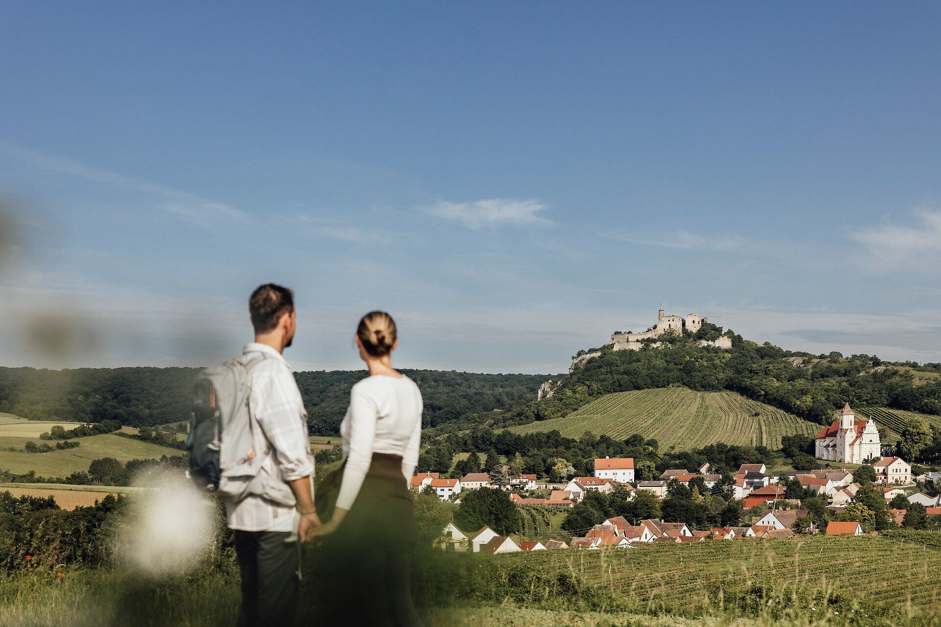 Ein romantisches Paar genießt die atemberaubende Aussicht auf die sanften Weinberge, die sich bis zur majestätischen Burgruine Falkenstein erstrecken. Die warmen Sonnenstrahlen und die frische Luft des Weinfrühlings schaffen eine einladende Atmosphäre für unvergessliche Erlebnisse in der Natur.