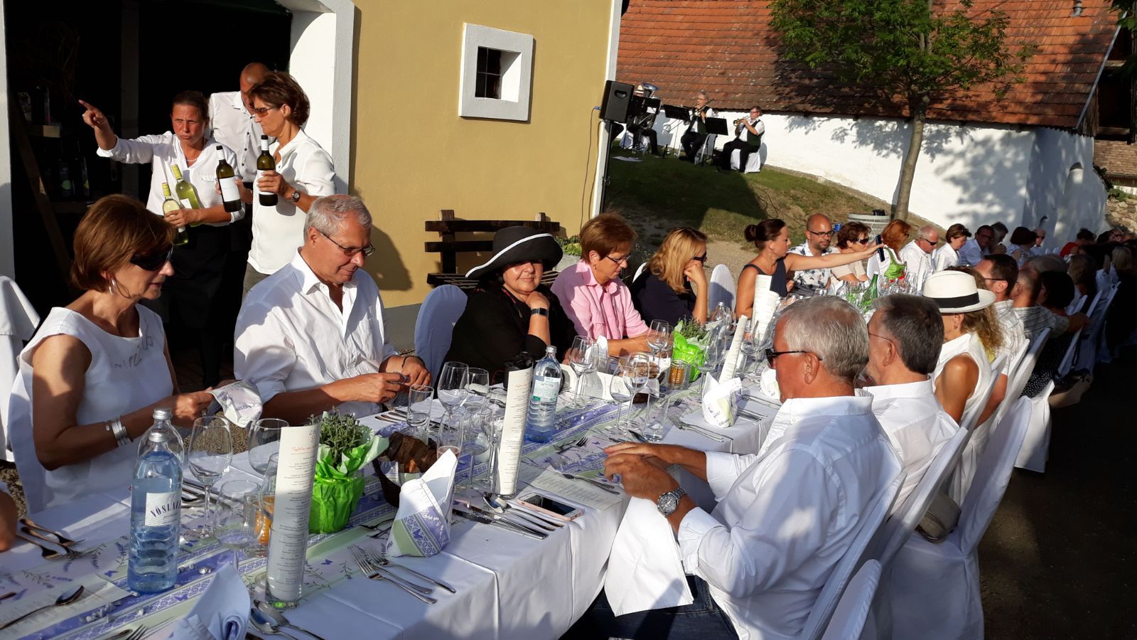 Menschen sitzen an einem langen Tisch im Freien, elegant gekleidet, mit Wein und Gläsern. Im Hintergrund spielt eine Band.