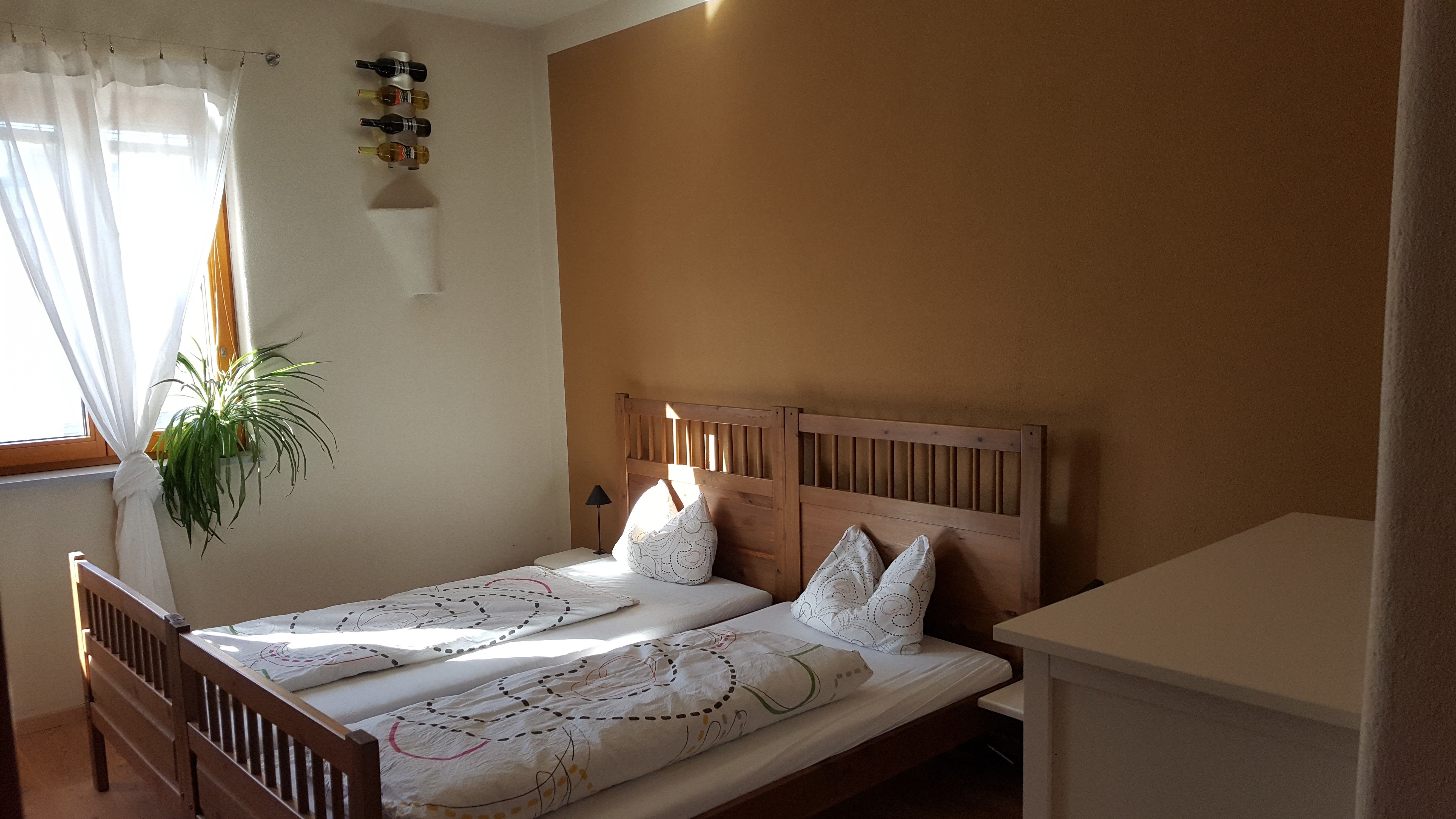 Ein gemütliches Schlafzimmer mit einem Doppelbett aus Holz, weißen Vorhängen, einer Pflanze am Fenster und einem Weinregal an der Wand.
