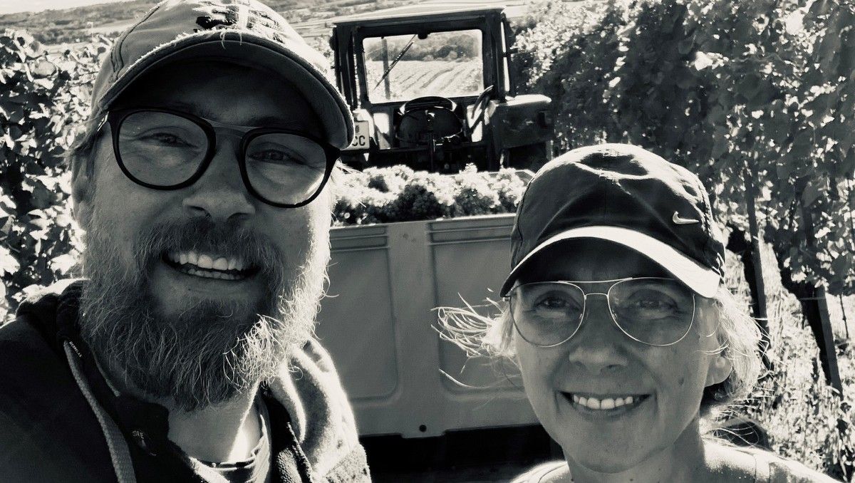 A winemaker couple smiles into the camera, a tractor with grapes in the background.