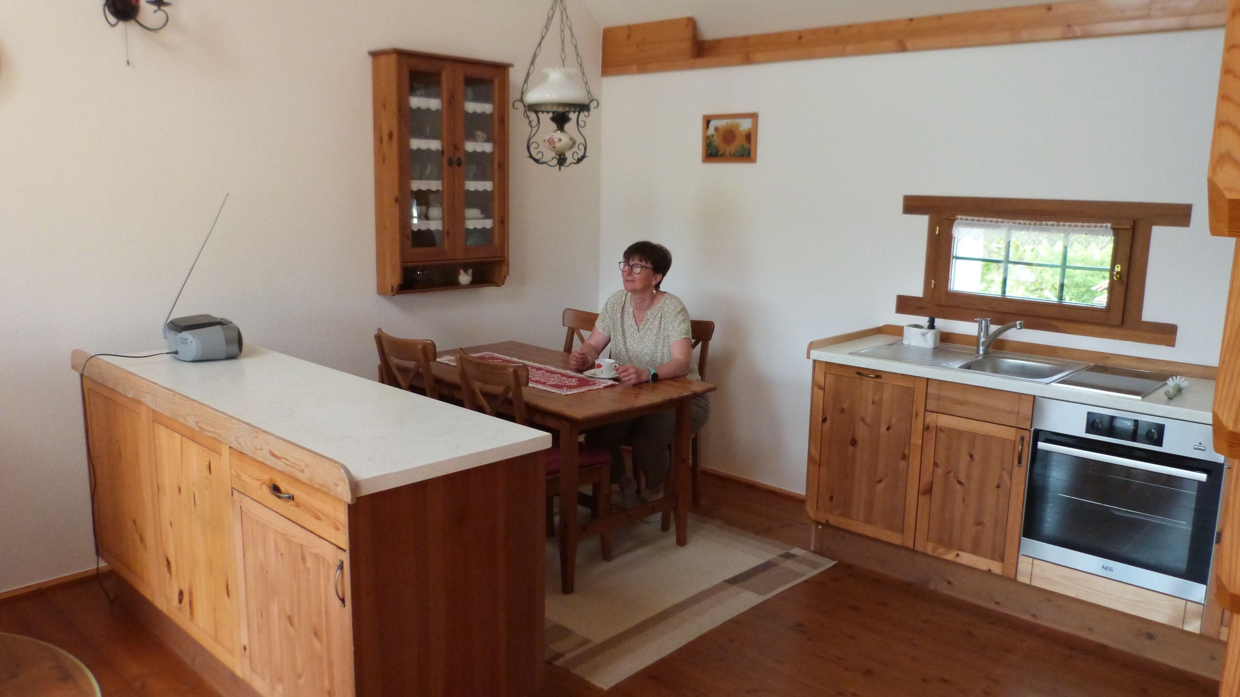 Eine Frau sitzt an einem Holztisch in einer gemütlichen Küche mit Holzschränken und einem kleinen Fenster.