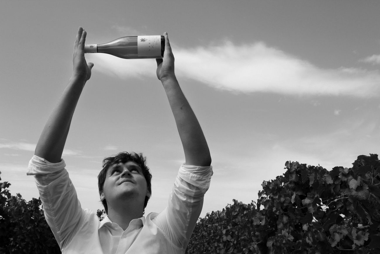 Person holding wine bottle in the air in a vineyard.