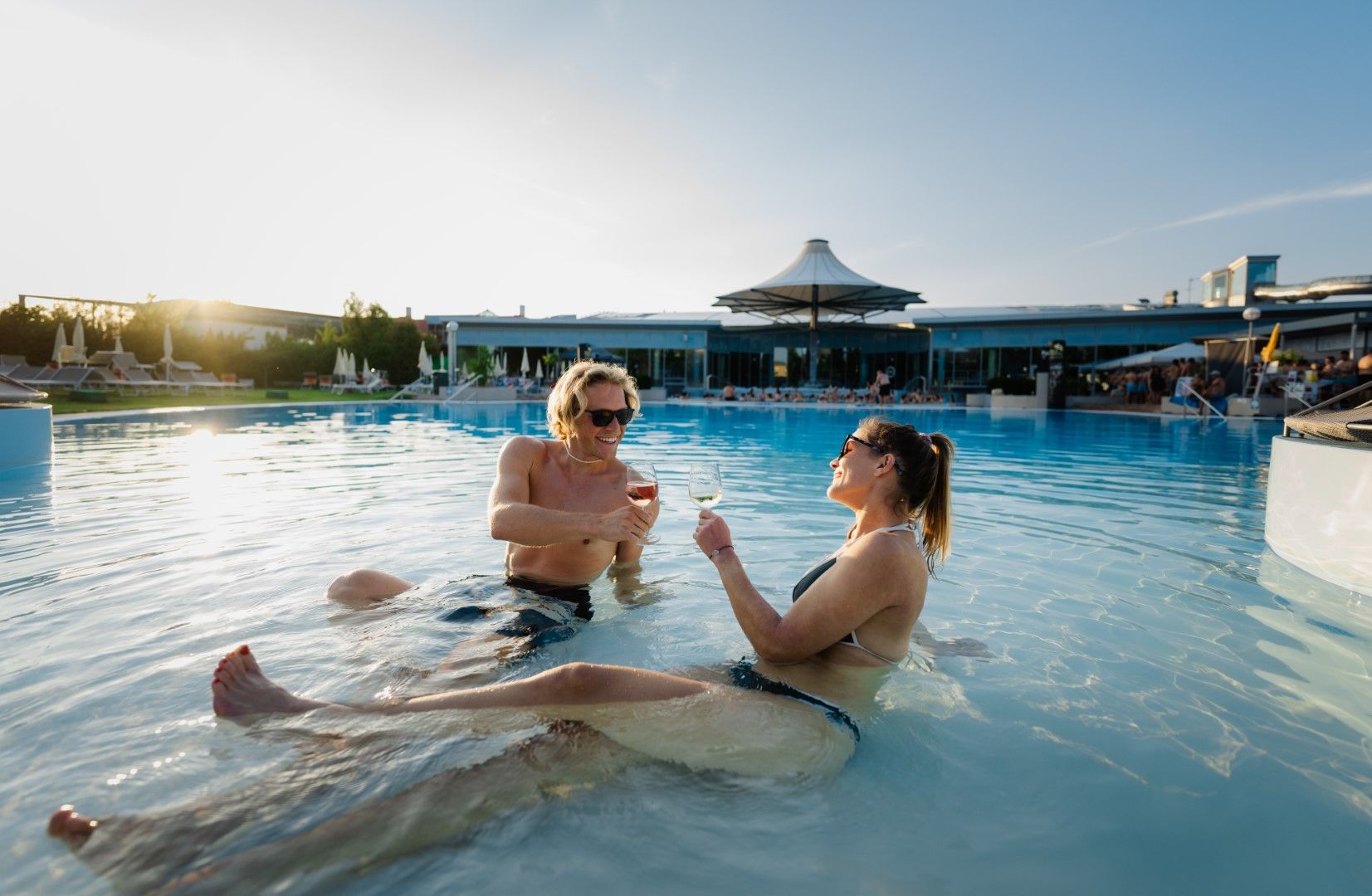 Two people relax in the pool at the Laa thermal spa resort and clink glasses of wine.