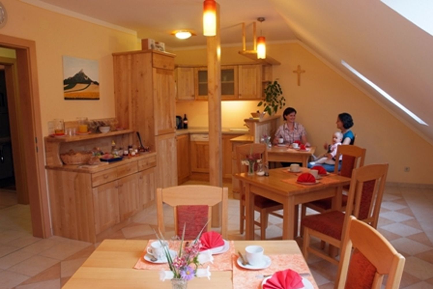 Ein gemütlicher Frühstücksraum mit Holzmöbeln, gedeckten Tischen und zwei Personen, die sich unterhalten. Ein Kind sitzt auf dem Schoß einer Person.