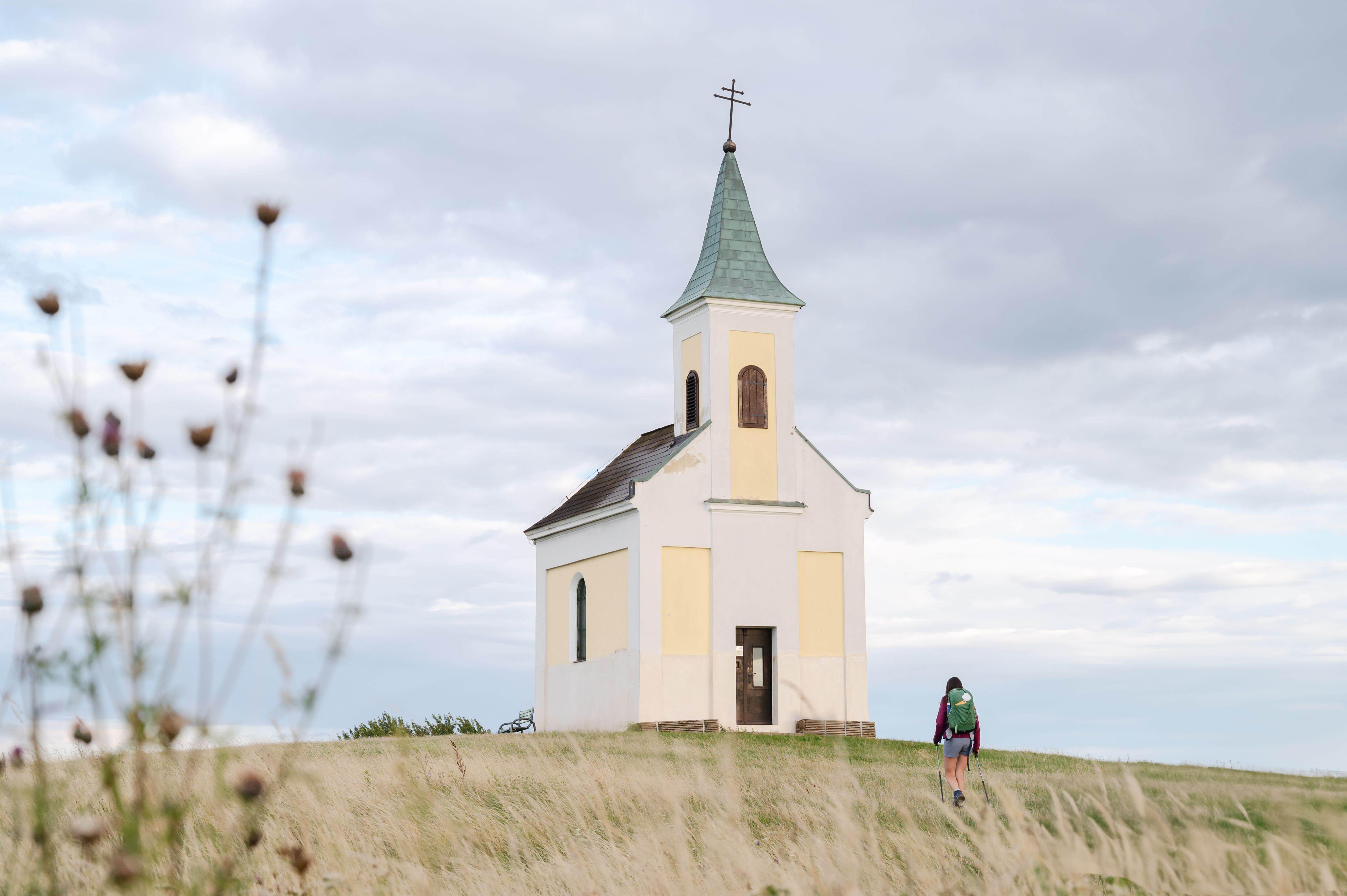 Eine kleine Kapelle auf einem Hügel mit einer Person, die darauf zugeht, umgeben von einer Wiese und bewölktem Himmel.