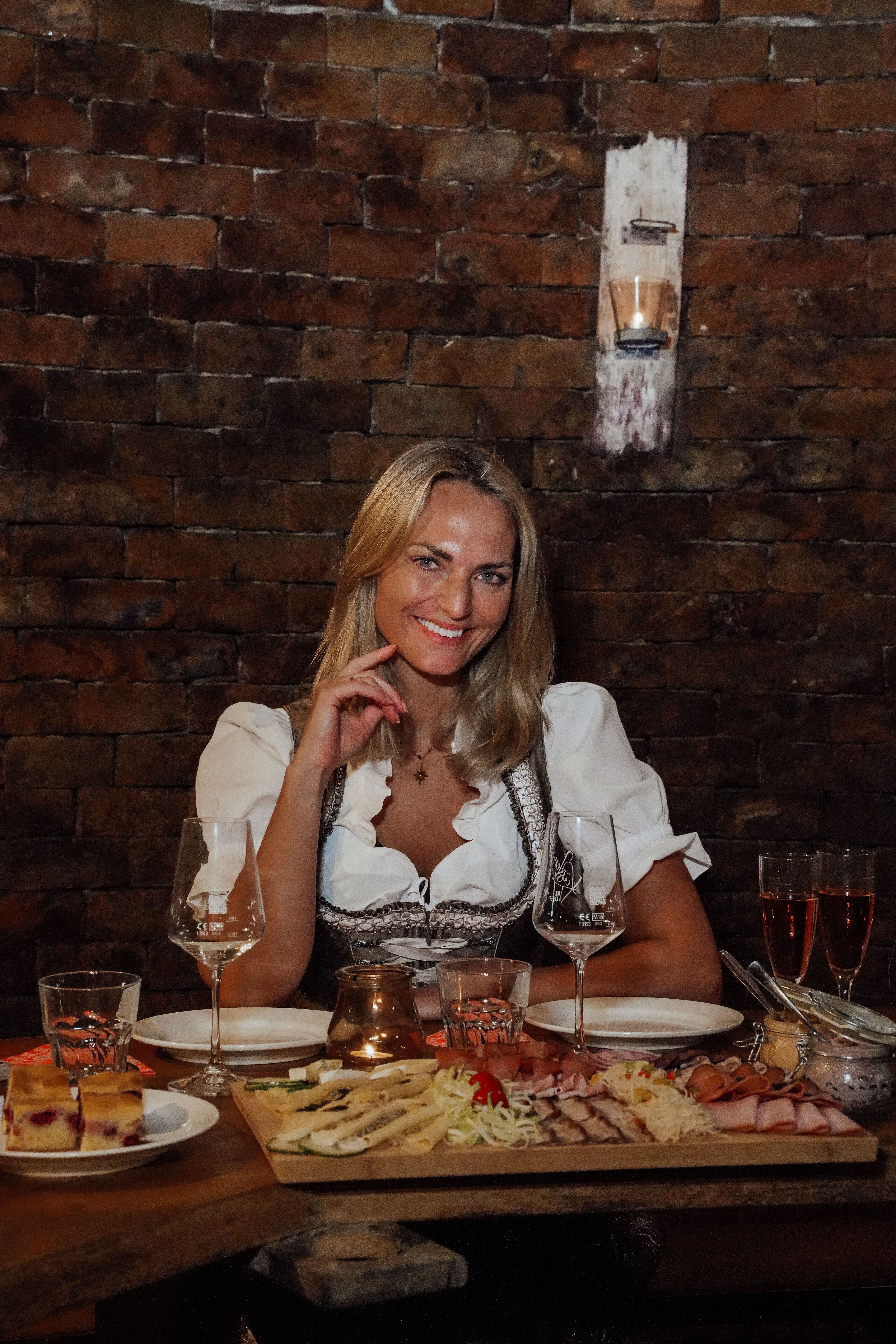 Eine lächelnde Frau sitzt im Winzerhof Küssler vor einer rustikalen Ziegelwand an einem Tisch mit Wein, Gläsern und einer Jause.