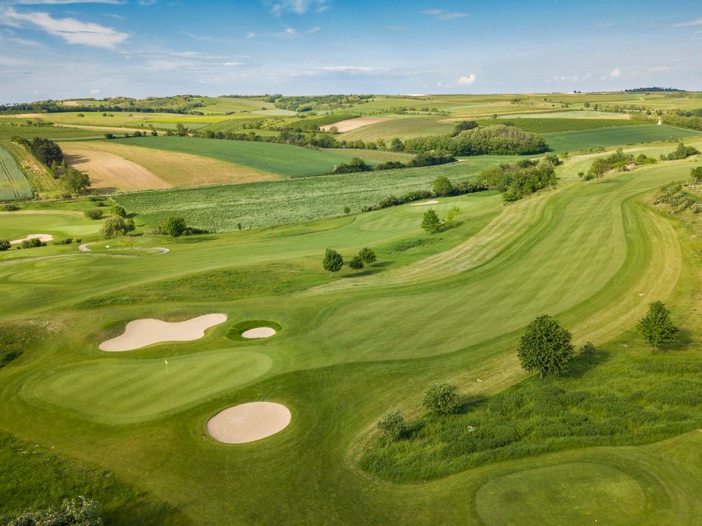 Luftaufnahme eines Golfplatzes mit grünen Fairways und Sandbunkern, umgeben von Feldern und Hügeln.