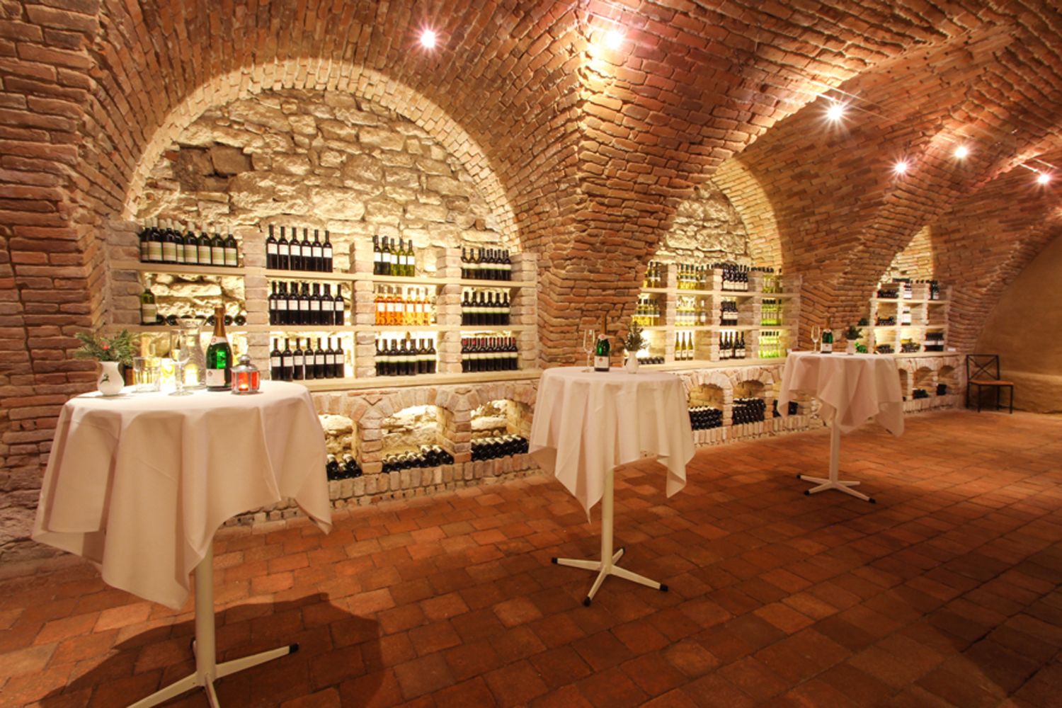Ein Weinkeller mit Backsteinwänden, Regalen voller Weinflaschen und Stehtischen mit weißen Tischdecken.