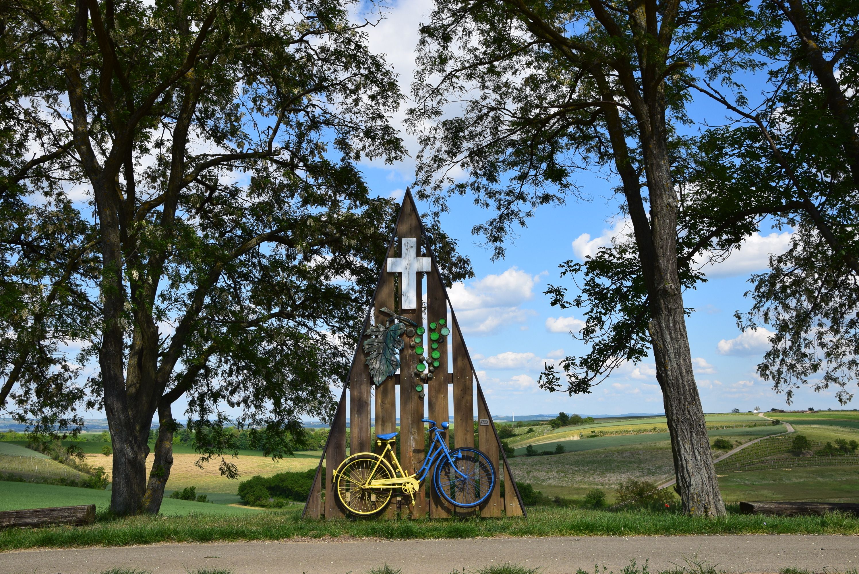 A shrine with a yellow and blue bicycle, surrounded by trees and fields under a blue sky.