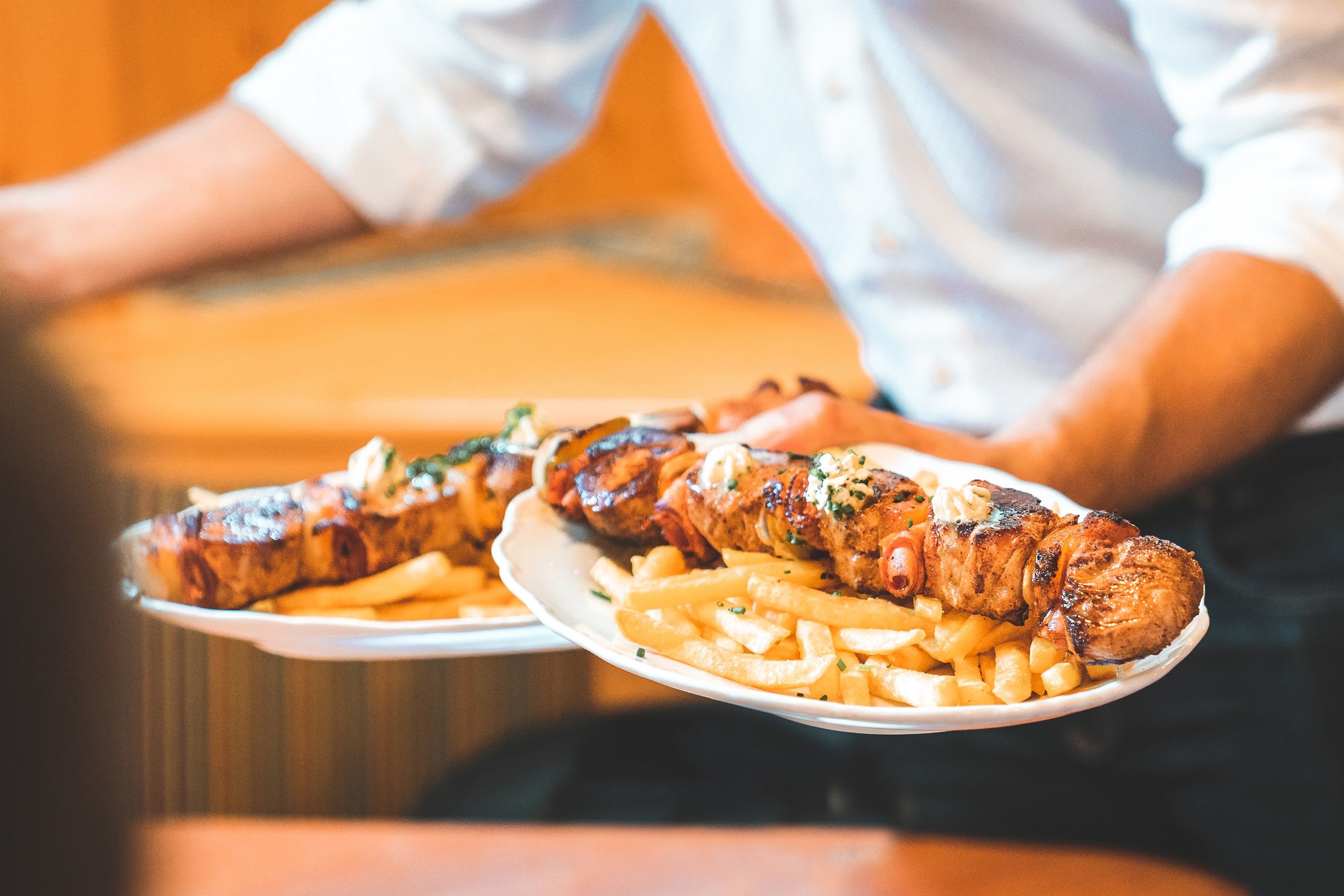 Ein Kellner serviert zwei Teller mit gegrillten Fleischspießen und Pommes frites.