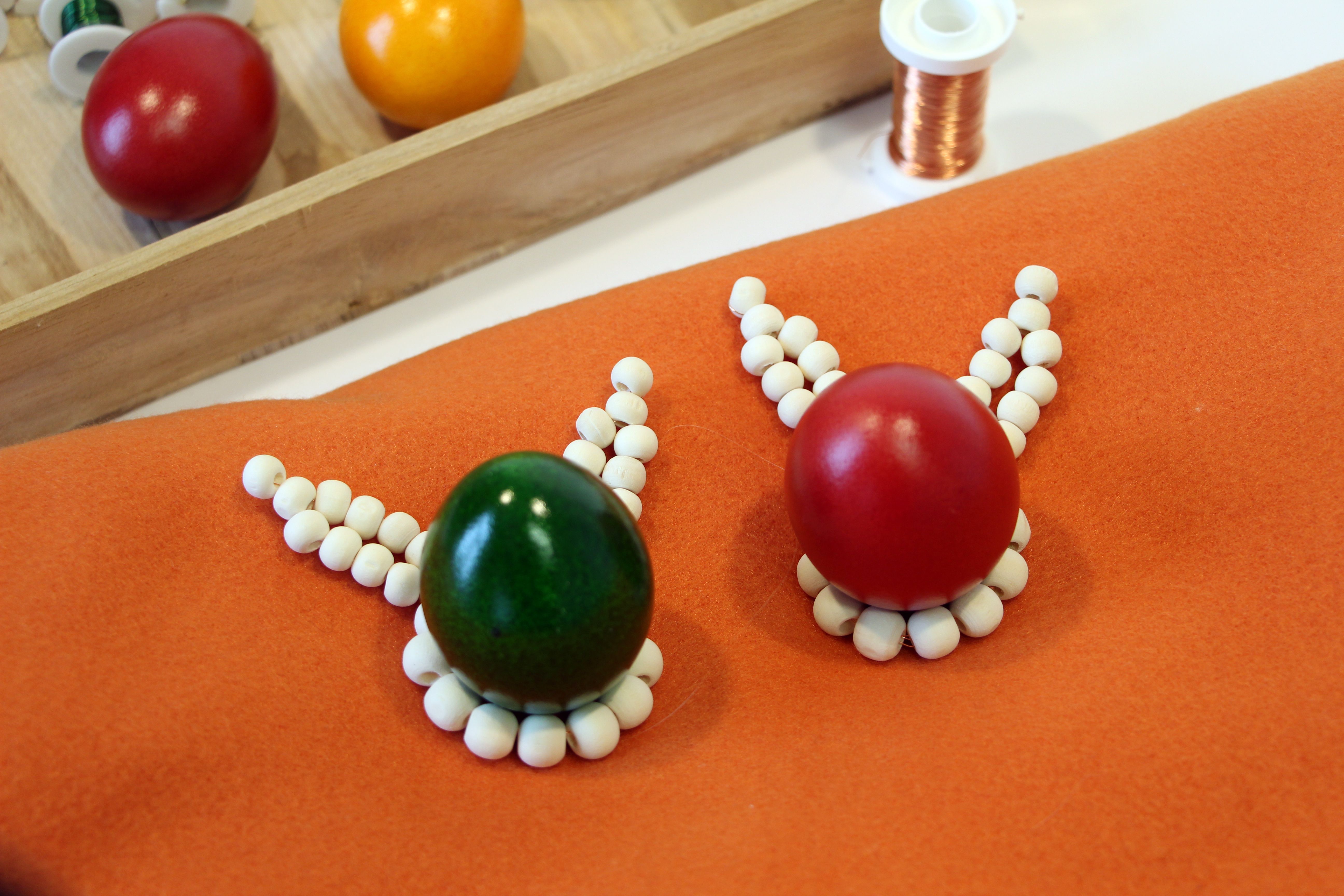 Zwei Eierbecher aus Holzperlen mit bunten Ostereiern sind auf einem Tisch platziert, im Hintergrund sieht man Eier und Bastelutensilien.