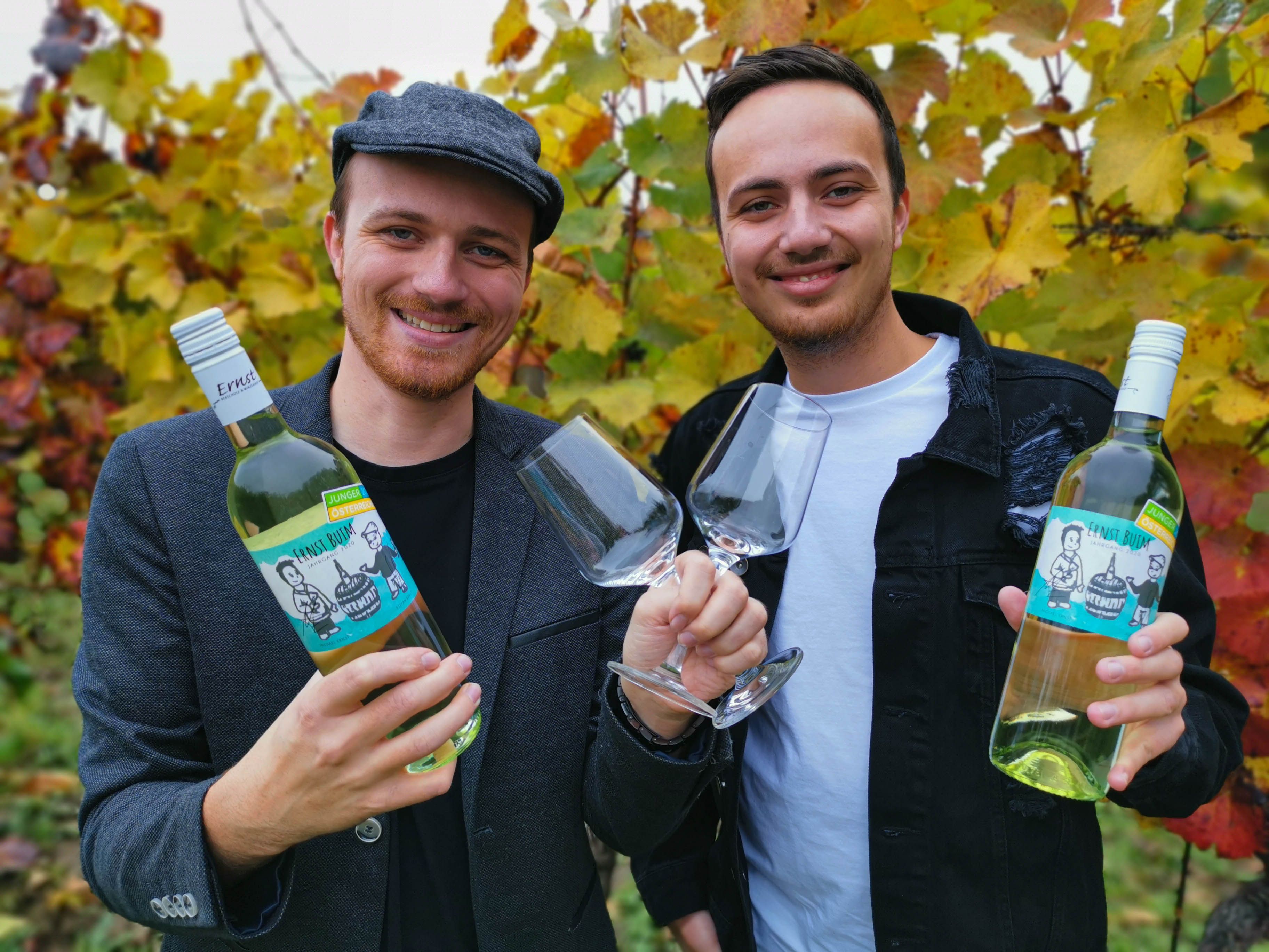Zwei Männer halten Weinflaschen und Gläser vor herbstlichen Weinreben.