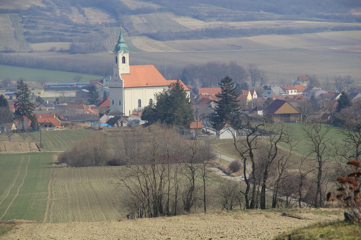 Landschaft mit Kirche und Dorf im Hintergrund, umgeben von Feldern und Bäumen.