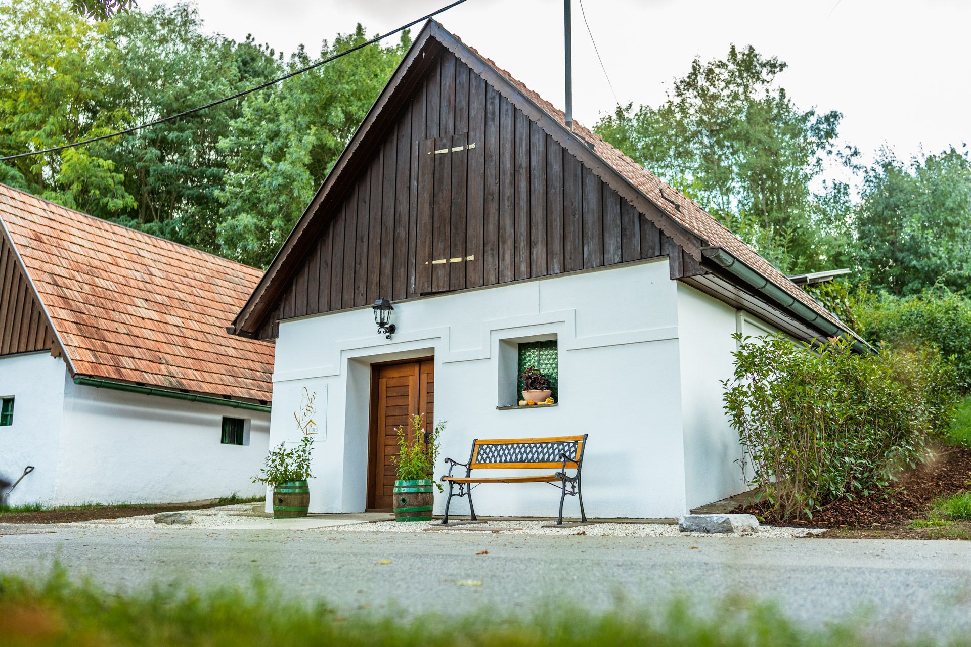 Winzerhof Küssler: weißes Gebäude mit dunklem Holzgiebel, rotem Ziegeldach, Bank und Pflanzen vor dem Eingang, umgeben von Grün.