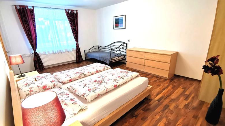 Schlafzimmer mit Doppelbett, Einzelbett, Kommode und Blumen in Vase.