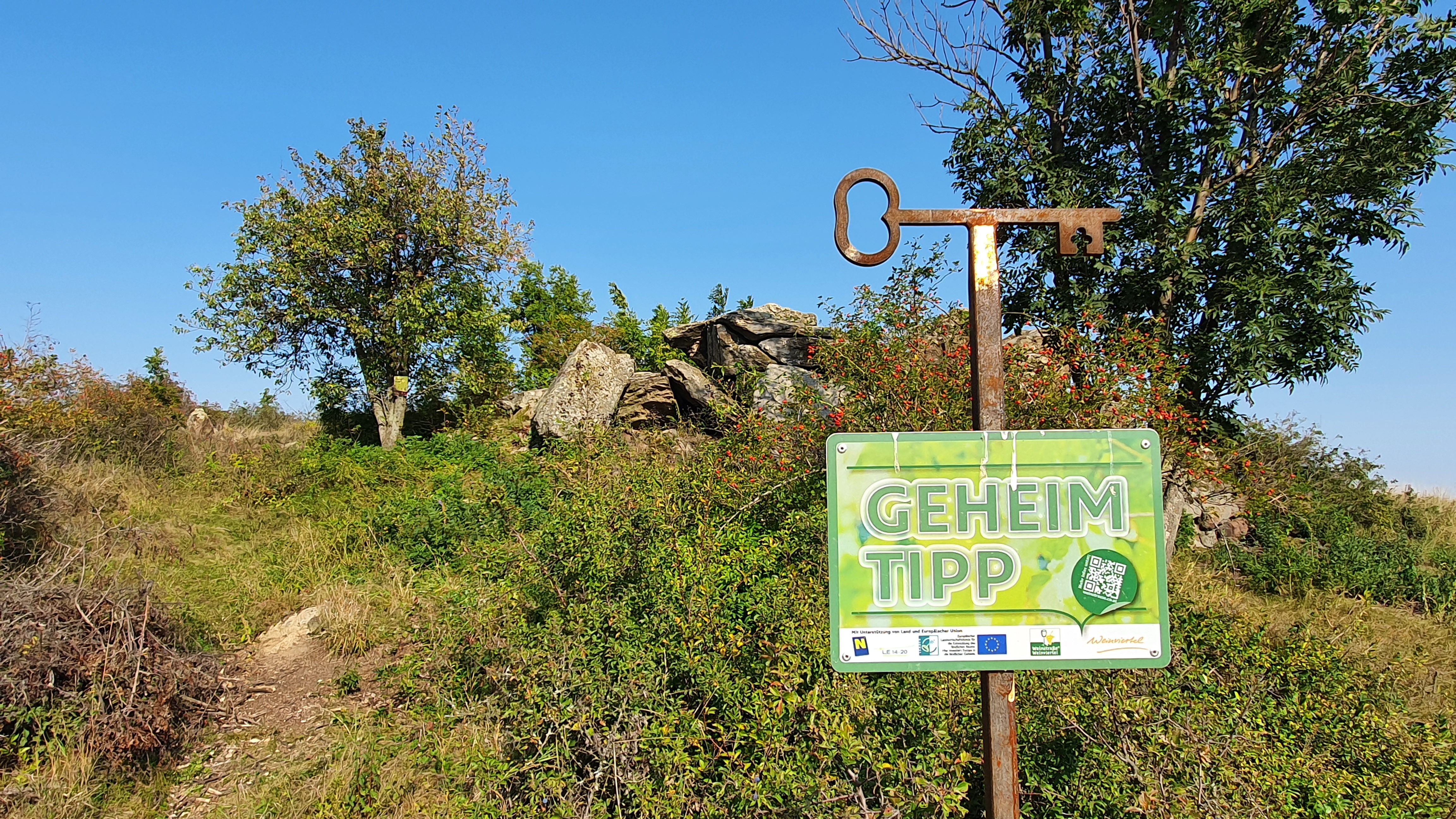 Ein Schild mit der Aufschrift 'Geheimtipp' vor einer natürlichen Landschaft mit Bäumen und Felsen.