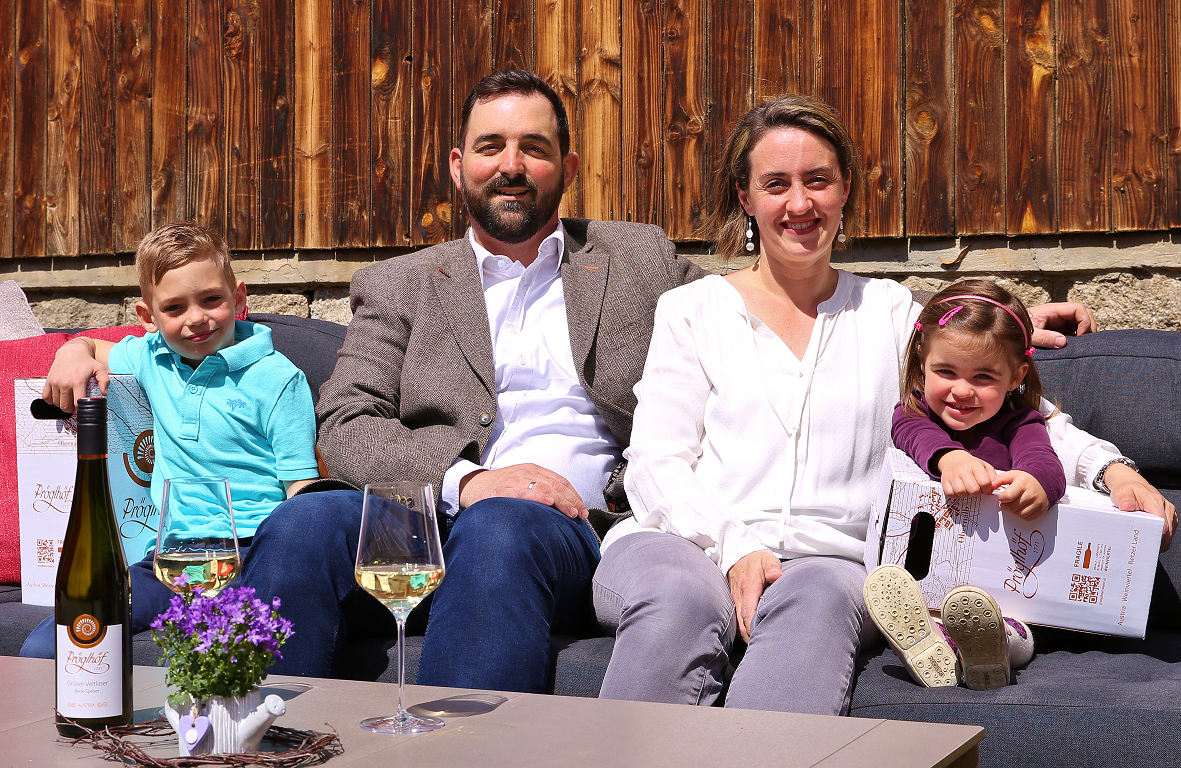 Eine Familie sitzt auf einem Sofa im Freien vor einer Holzwand, mit Weinflaschen und Gläsern auf dem Tisch.