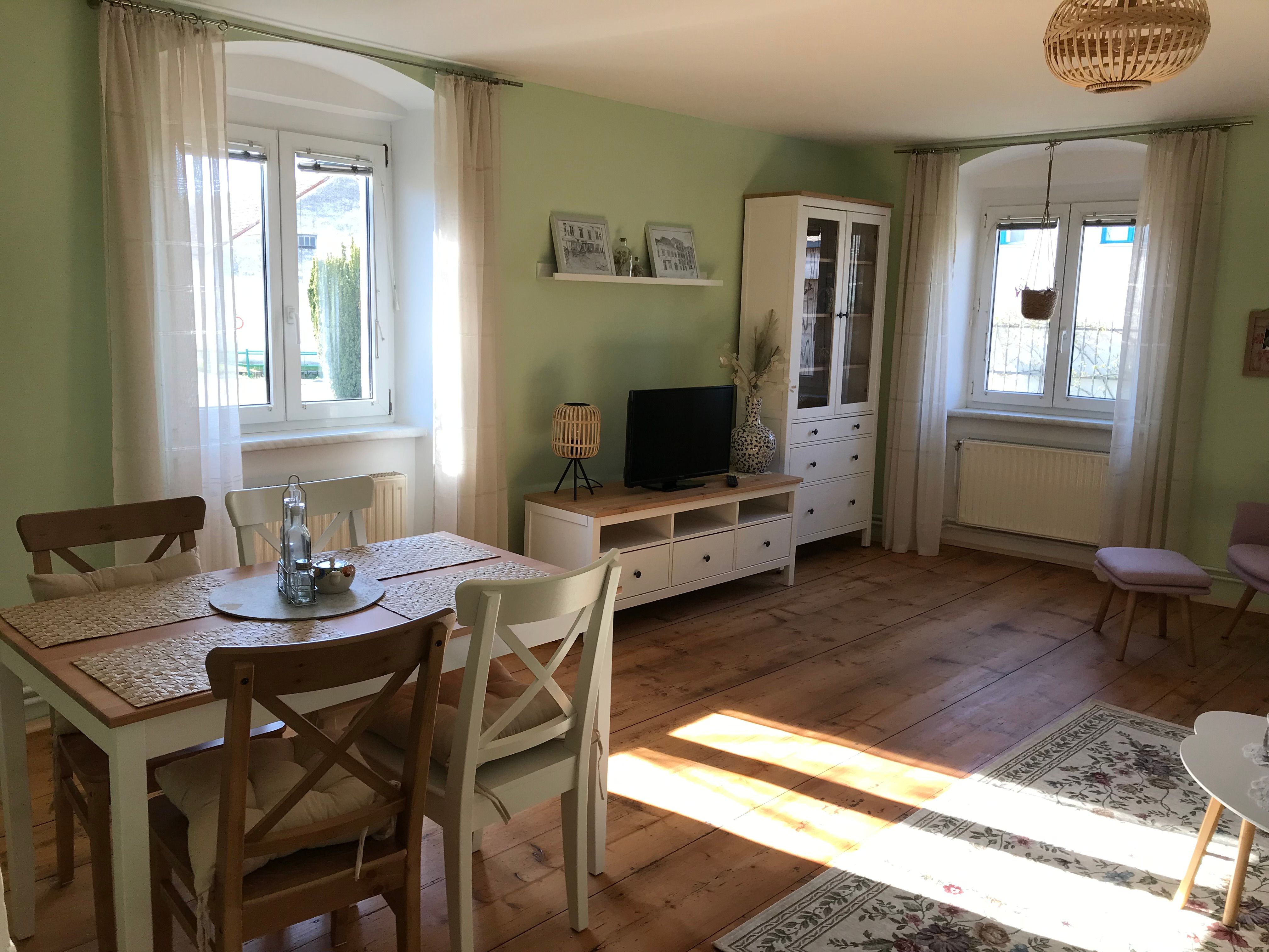 Bright room with dining table, chairs, TV cabinet and large windows.