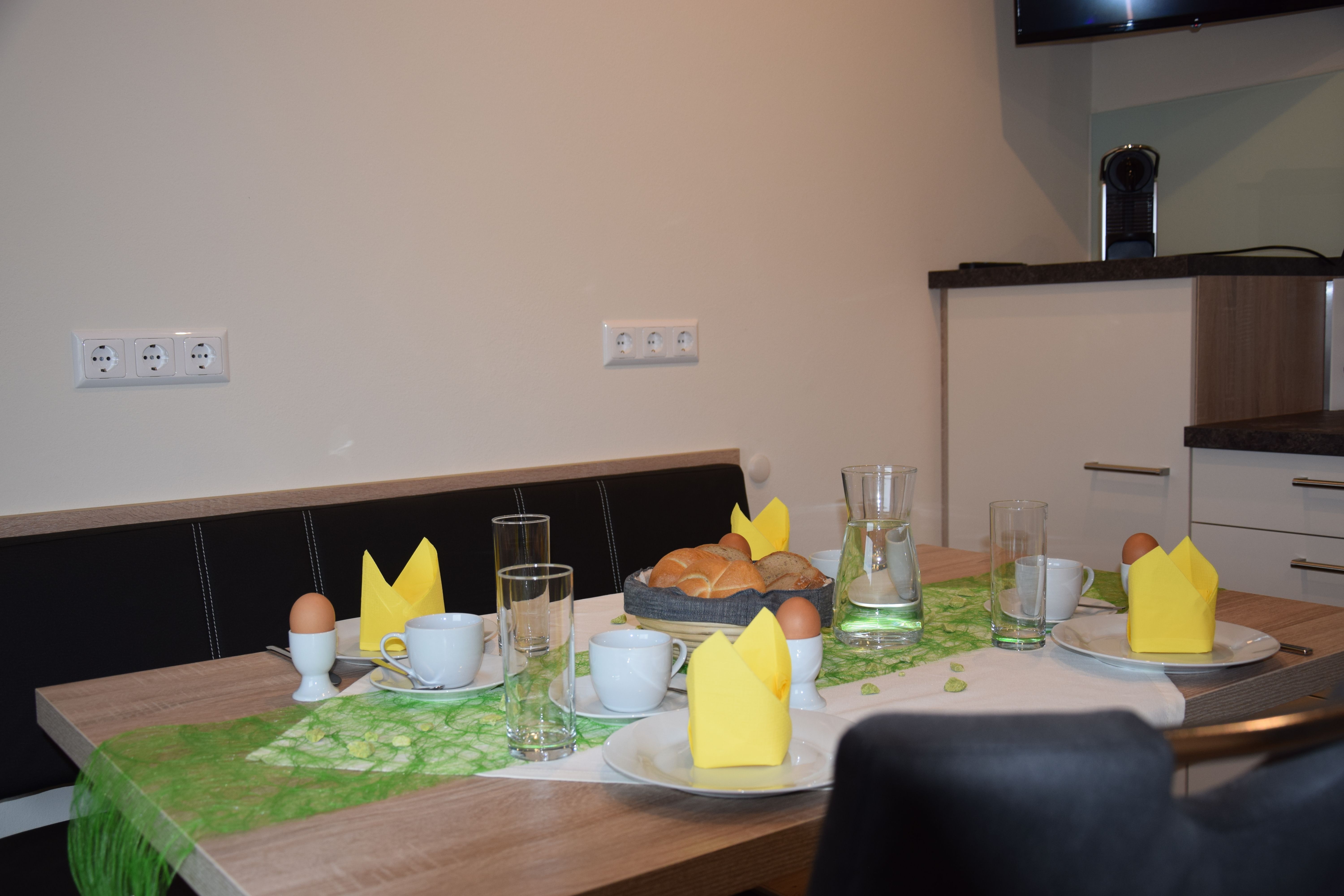 Frühstückstisch mit Brötchen, Eiern, Tassen und gelben Servietten.