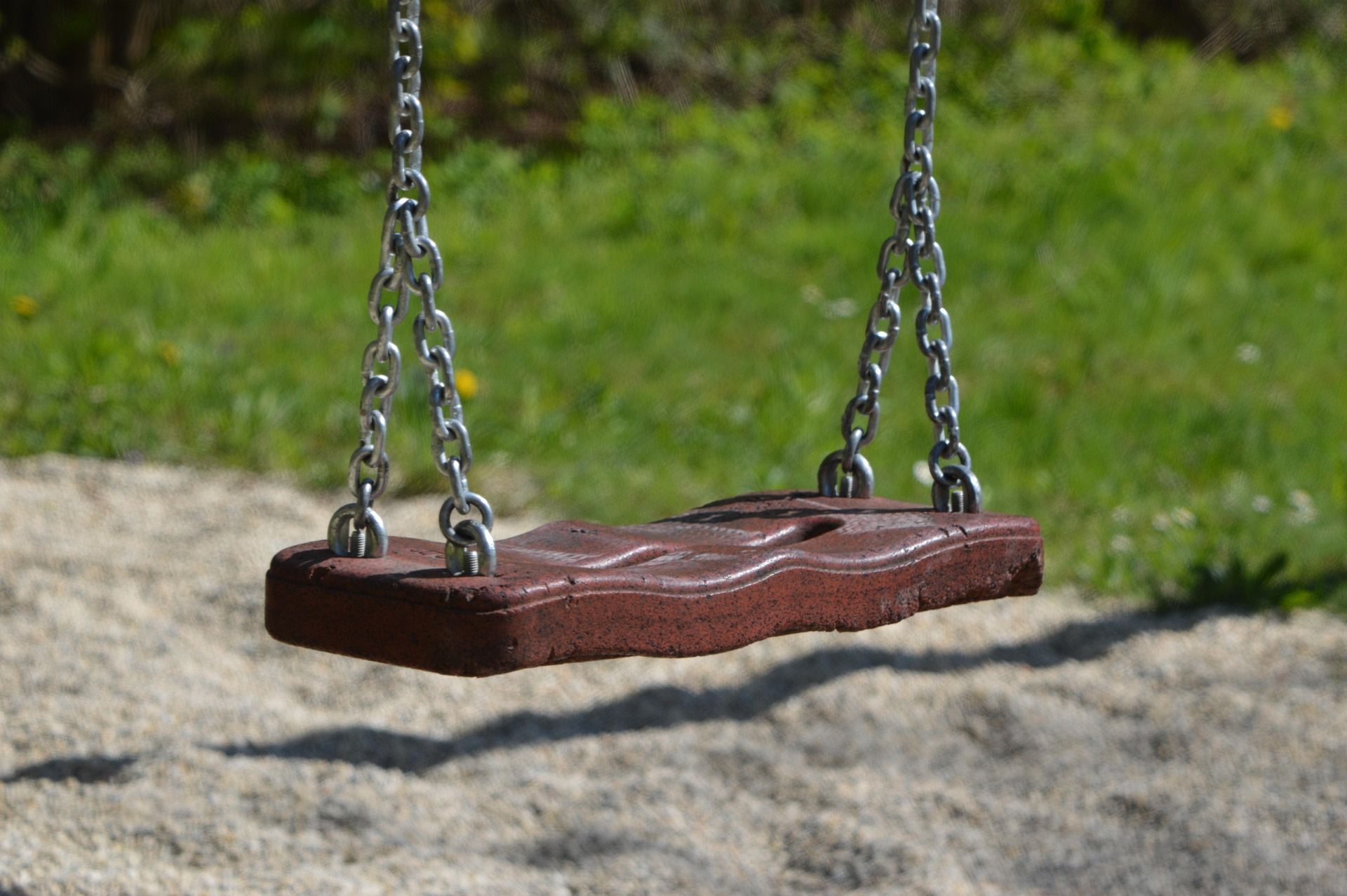 An empty swing made of wood and metal chains hangs over a gravel floor, surrounded by green grass.