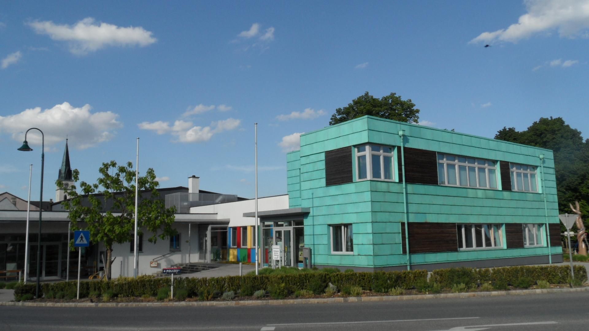 Modernes Gebäude mit türkisfarbener Fassade in Guntersdorf, daneben ein Kirchturm im Hintergrund.