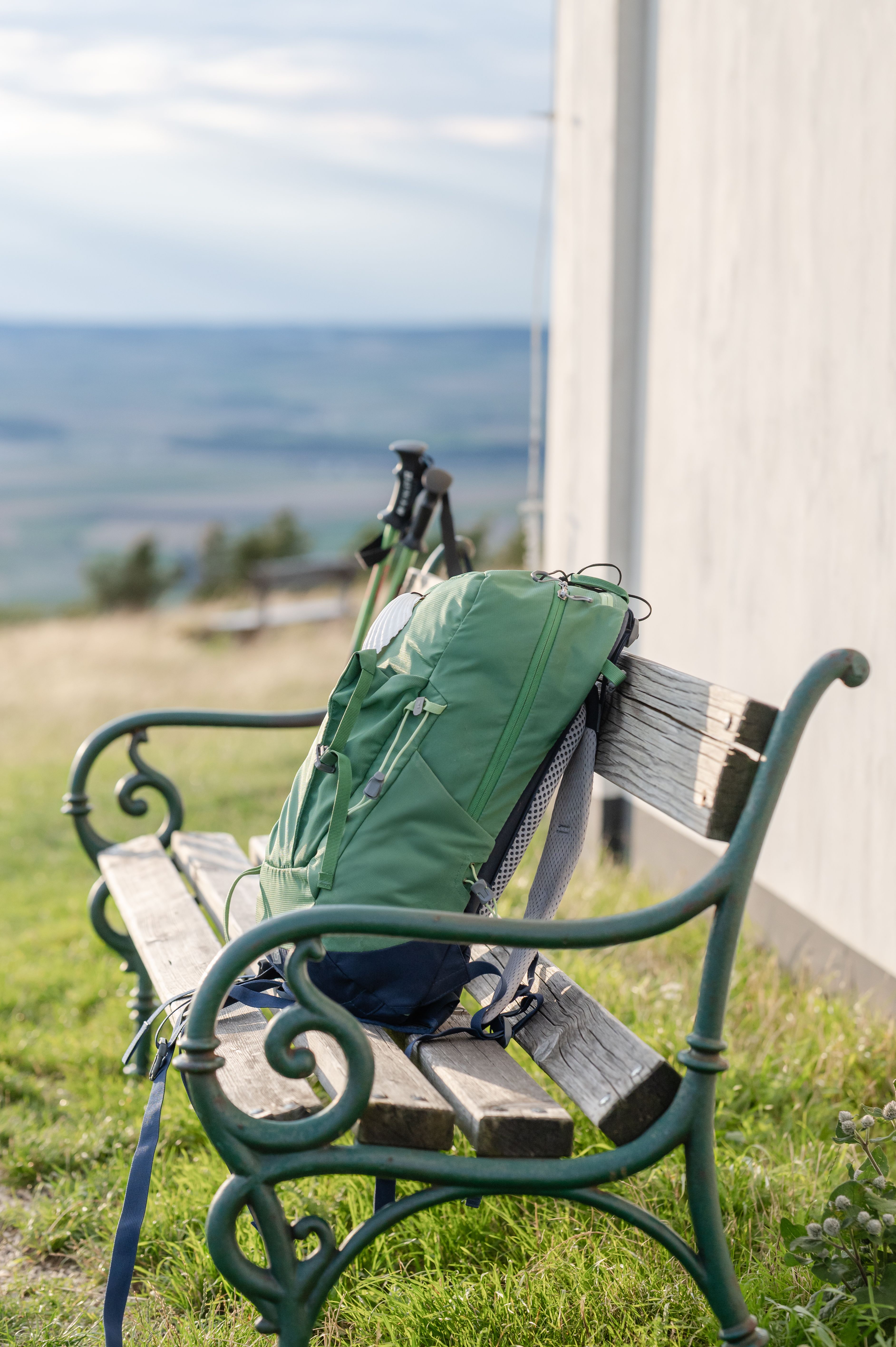 Ein grüner Rucksack liegt auf einer Holzbank mit Metallverzierungen, im Hintergrund eine weite Landschaft.