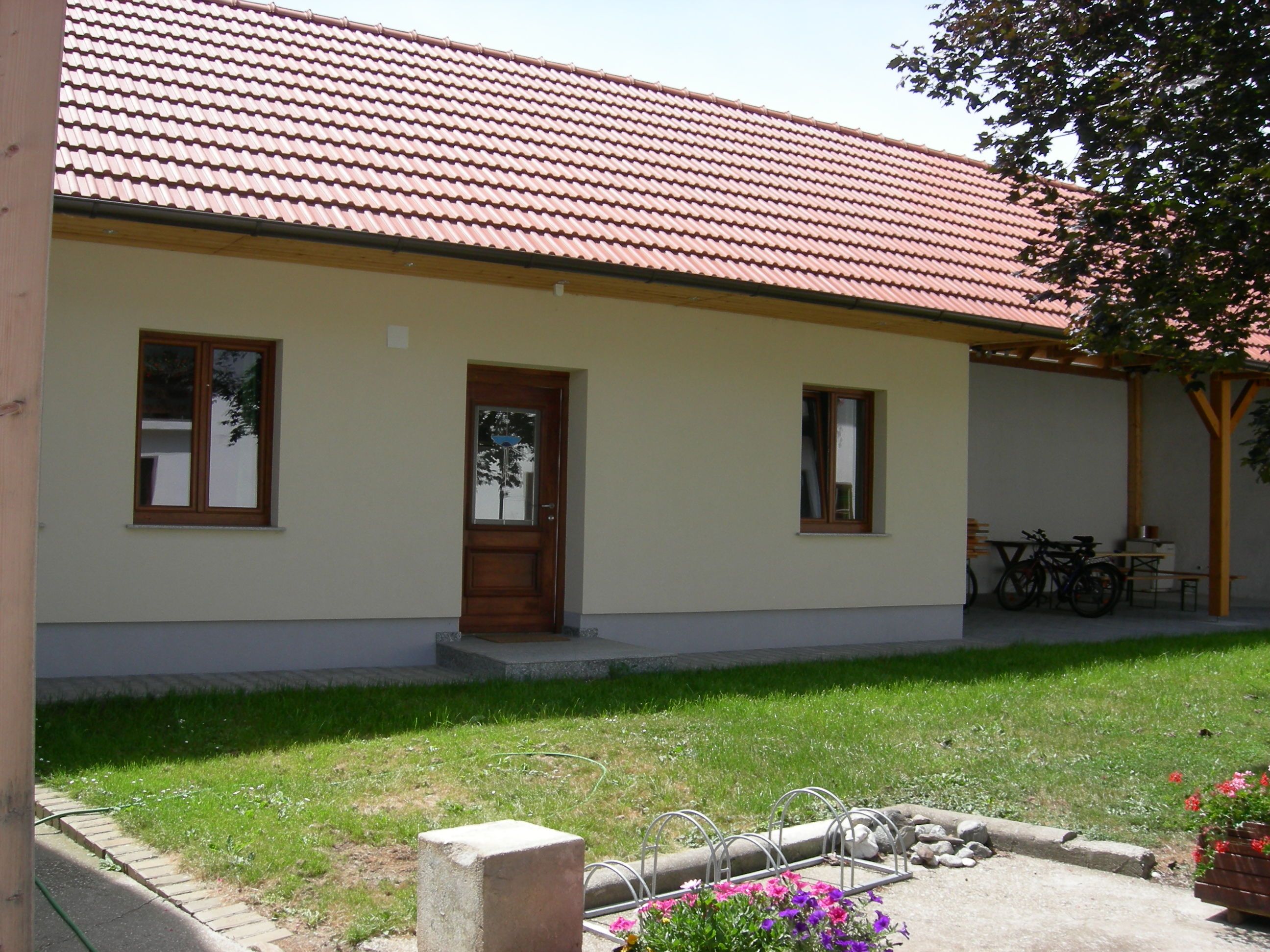 Ein Innenhof mit einem Gebäude mit rotem Ziegeldach, Holztür und Fenstern. Im Vordergrund sind Blumen und ein Fahrradständer zu sehen.